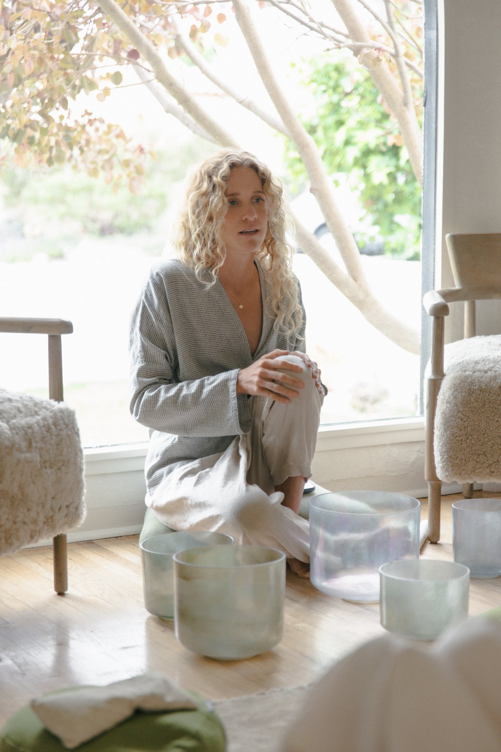A woman with curly blonde hair sitting by a large window, surrounded by glass candle holders on a wooden floor, with greenery visible outside.