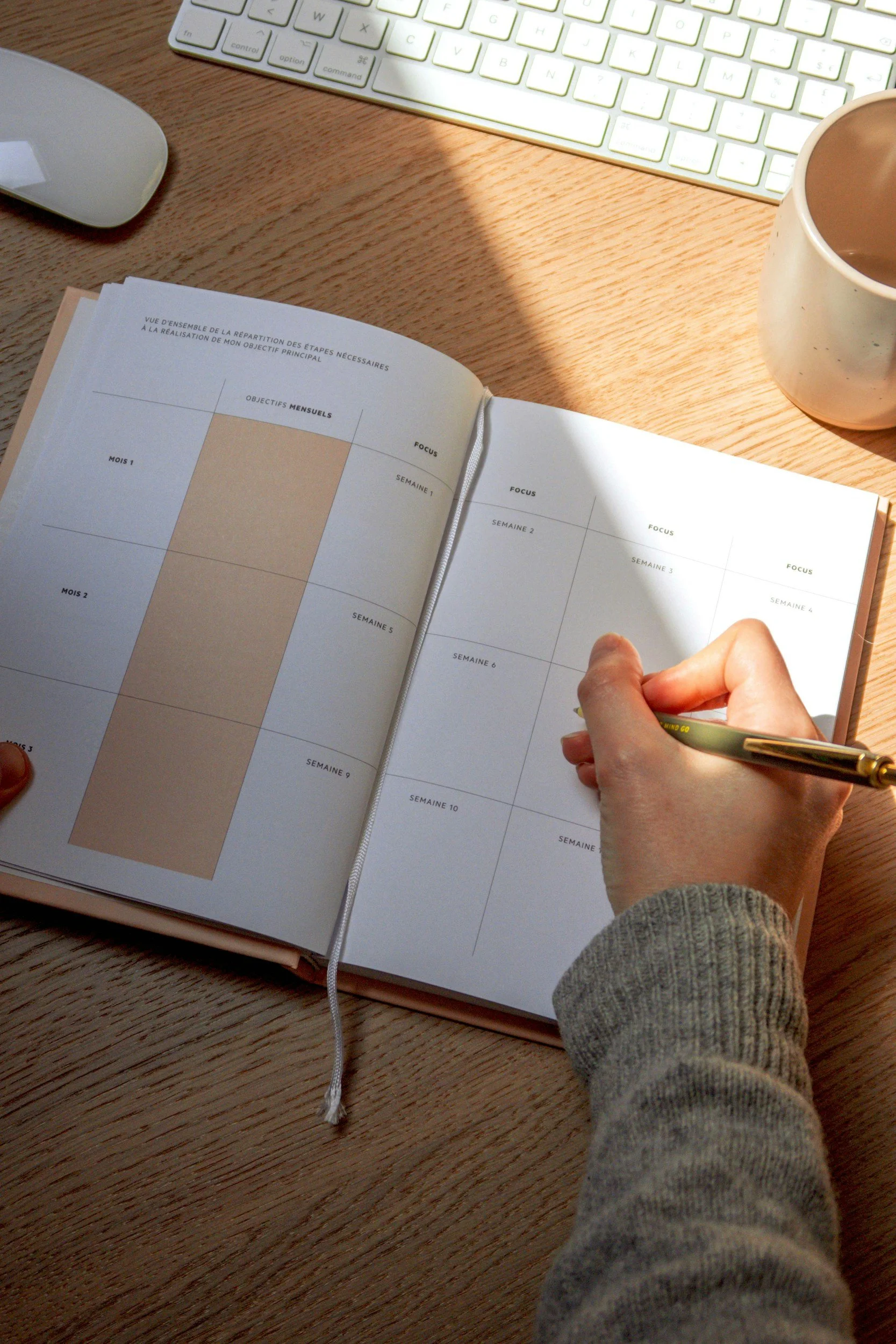 An open planner or journal on a wooden desk with a person writing in it with a gold pen. Surrounding items include a white wireless mouse, a white keyboard with backlit keys, and a beige mug filled with coffee or tea.