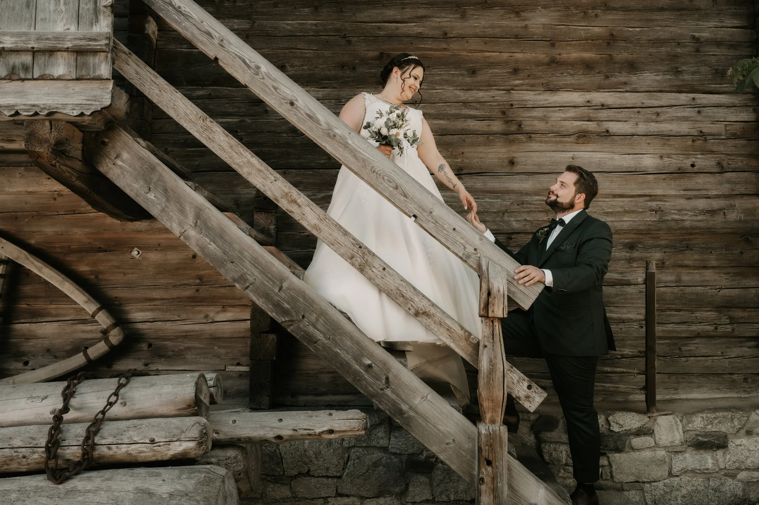 A bride in a white wedding dress holding a bouquet, smiling and looking at her groom, who is dressed in a black tuxedo, as he kneels on one knee on wooden stairs outside a rustic wooden building.