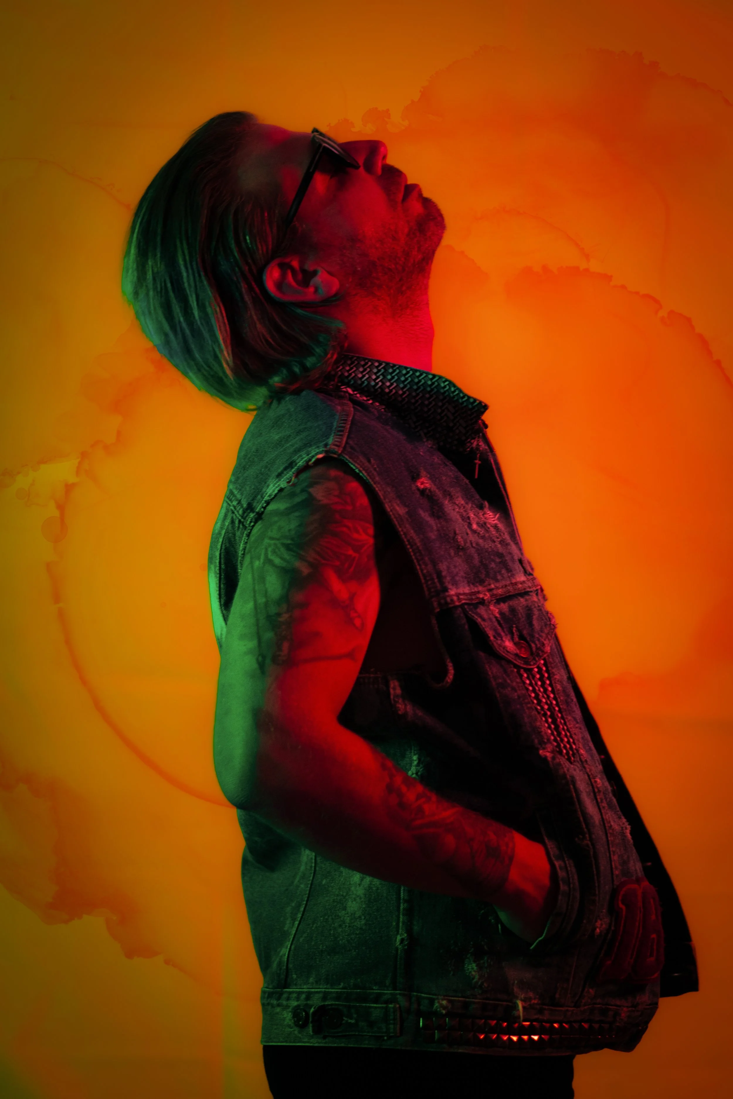 Side view of a man with long hair, beard, wearing glasses, a sleeveless denim vest with studs, standing against an orange background.