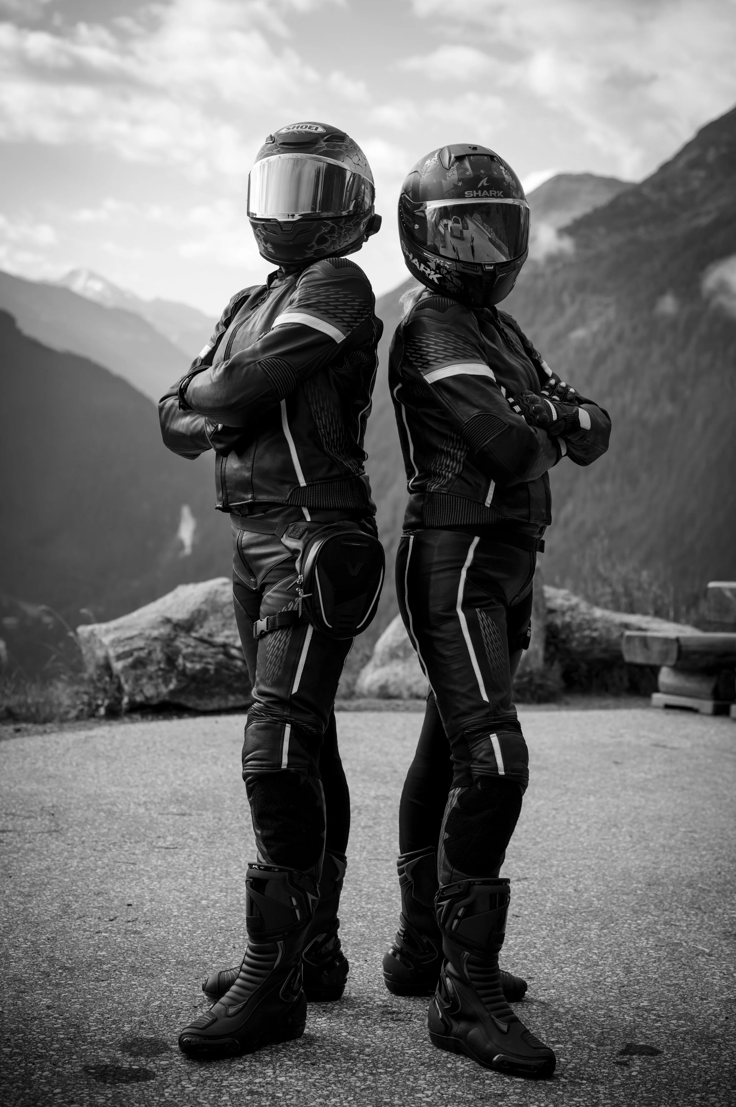 Two motorcyclists in full gear, including helmets, jackets, pants, and boots, standing back to back outdoors with mountains and cloudy sky in the background.