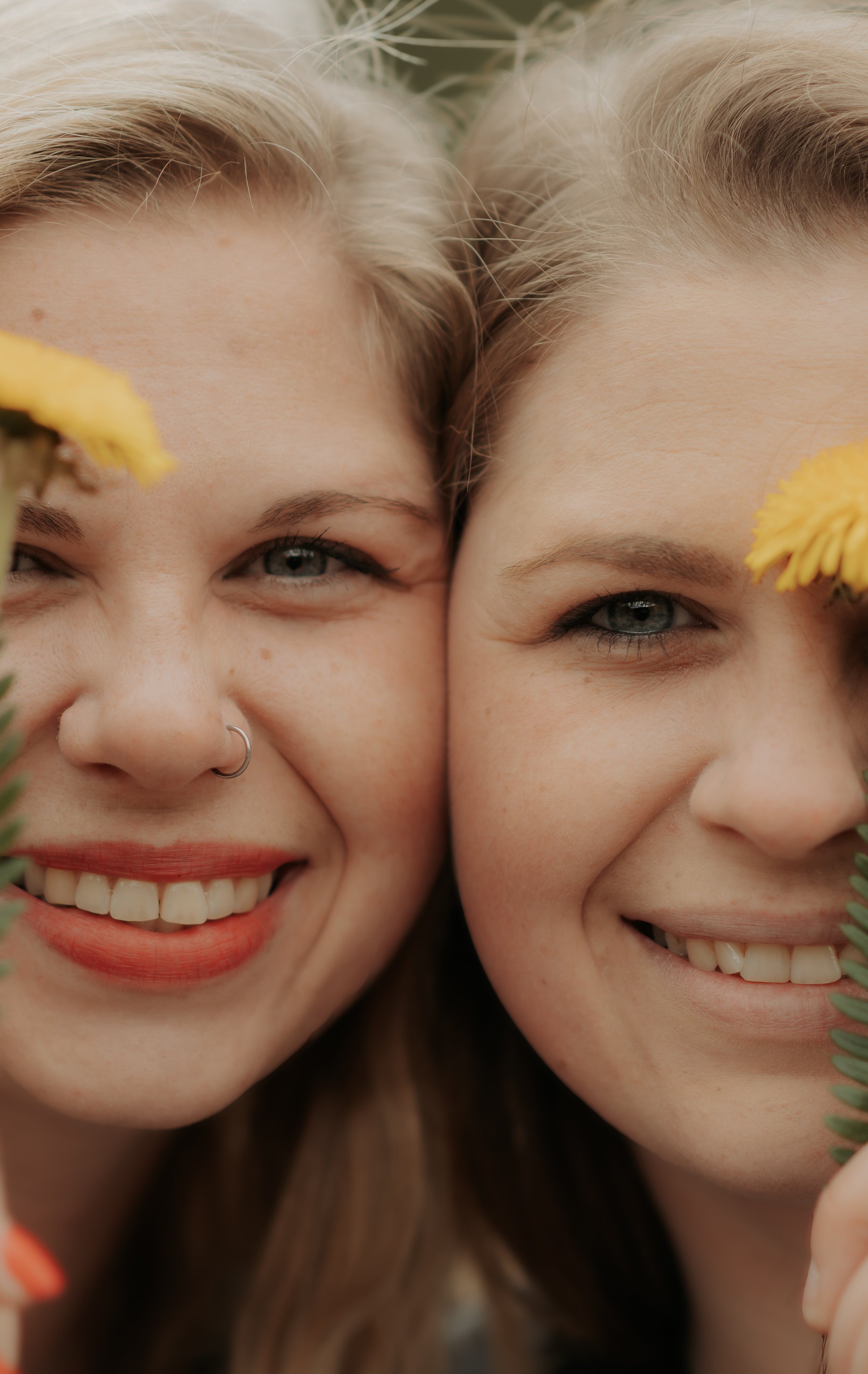 Close-up of two smiling young women with fair skin and blonde hair, partially obscured by yellow flowers in the foreground.