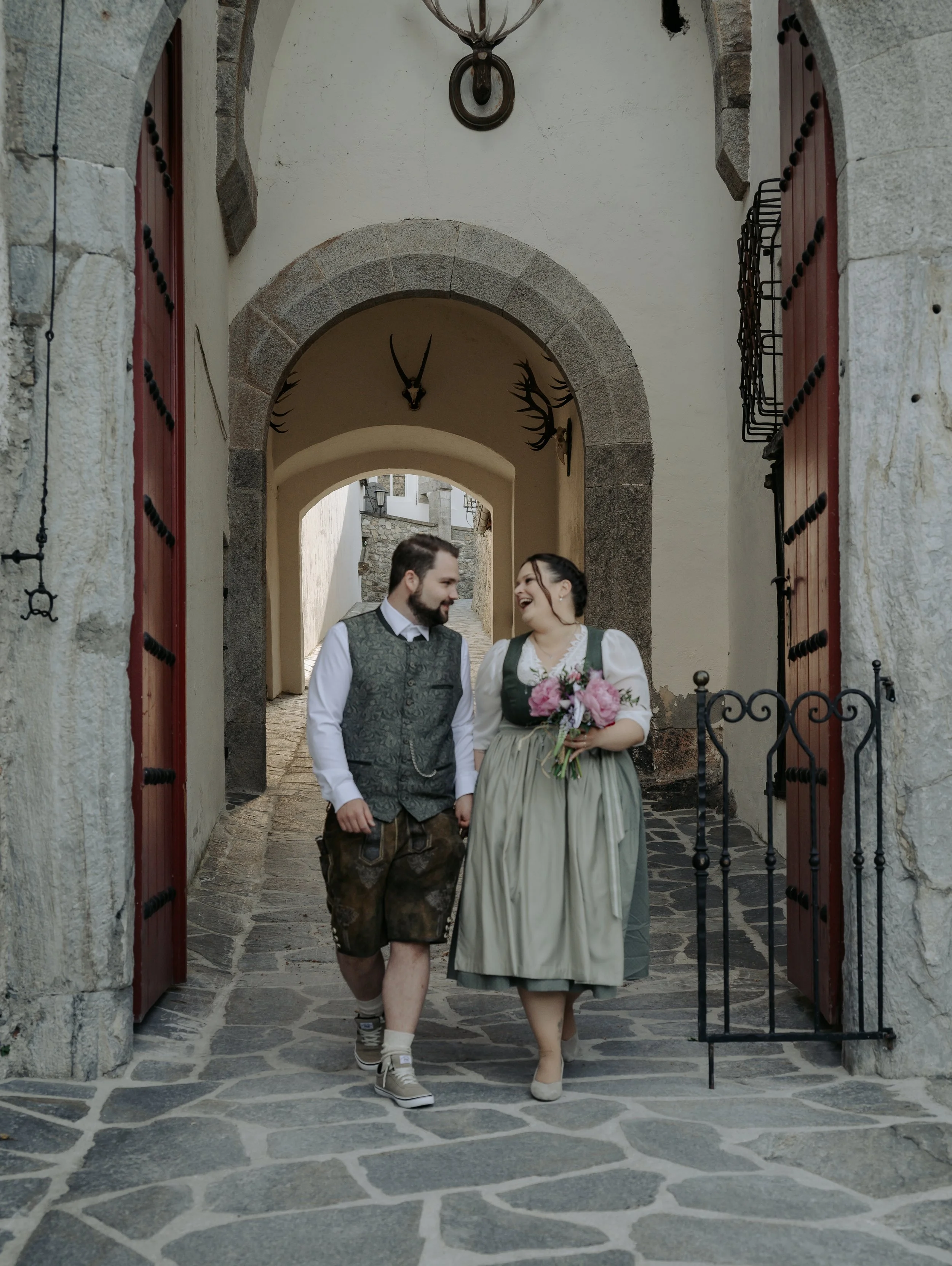 A smiling couple dressed in traditional Bavarian clothing walking through a stone archway in a European village, with deer antler and animal skull decorations on the wall.