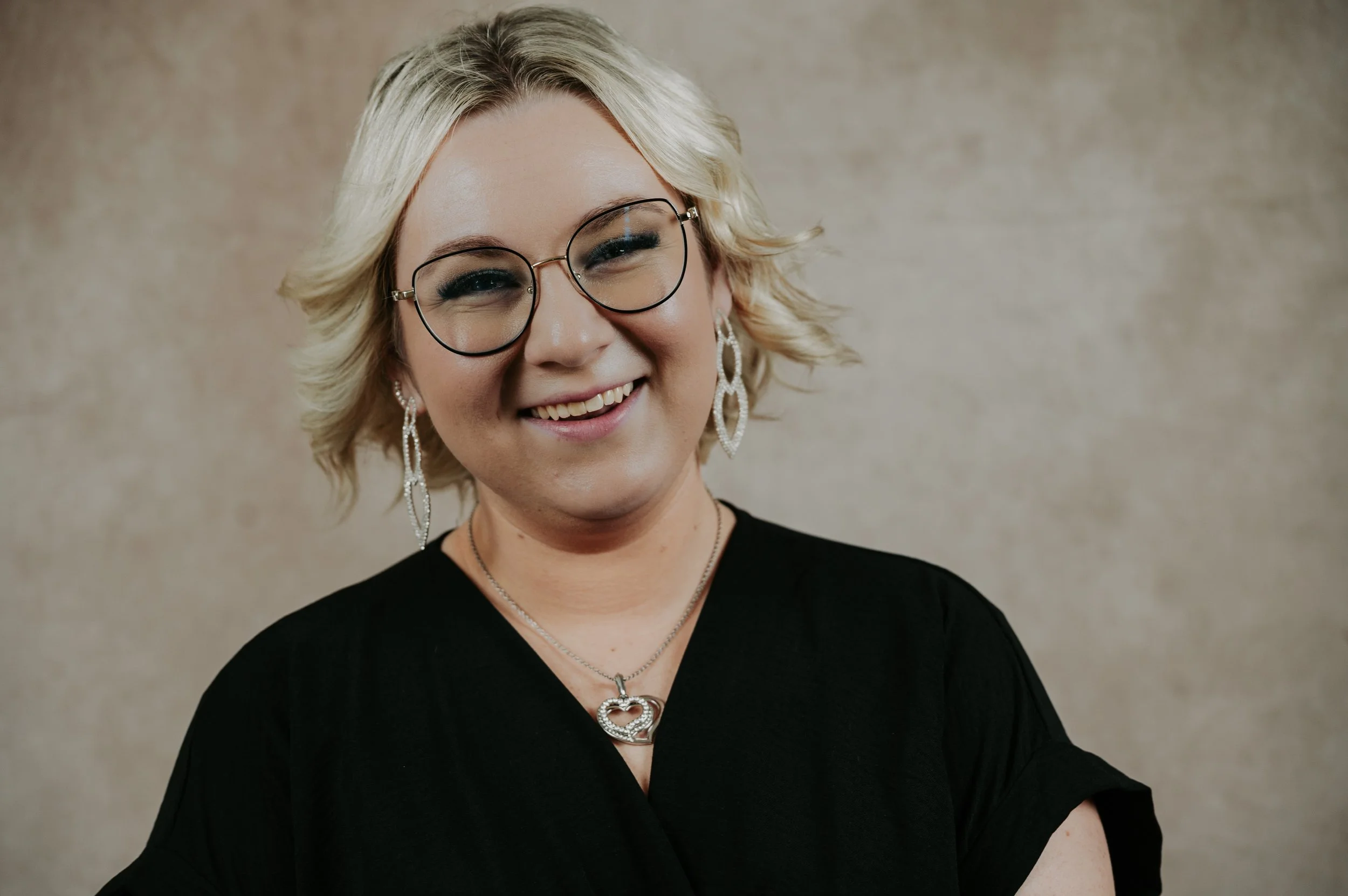 A woman with blond hair, wearing glasses, earrings, and a black top, smiling at the camera against a neutral background.