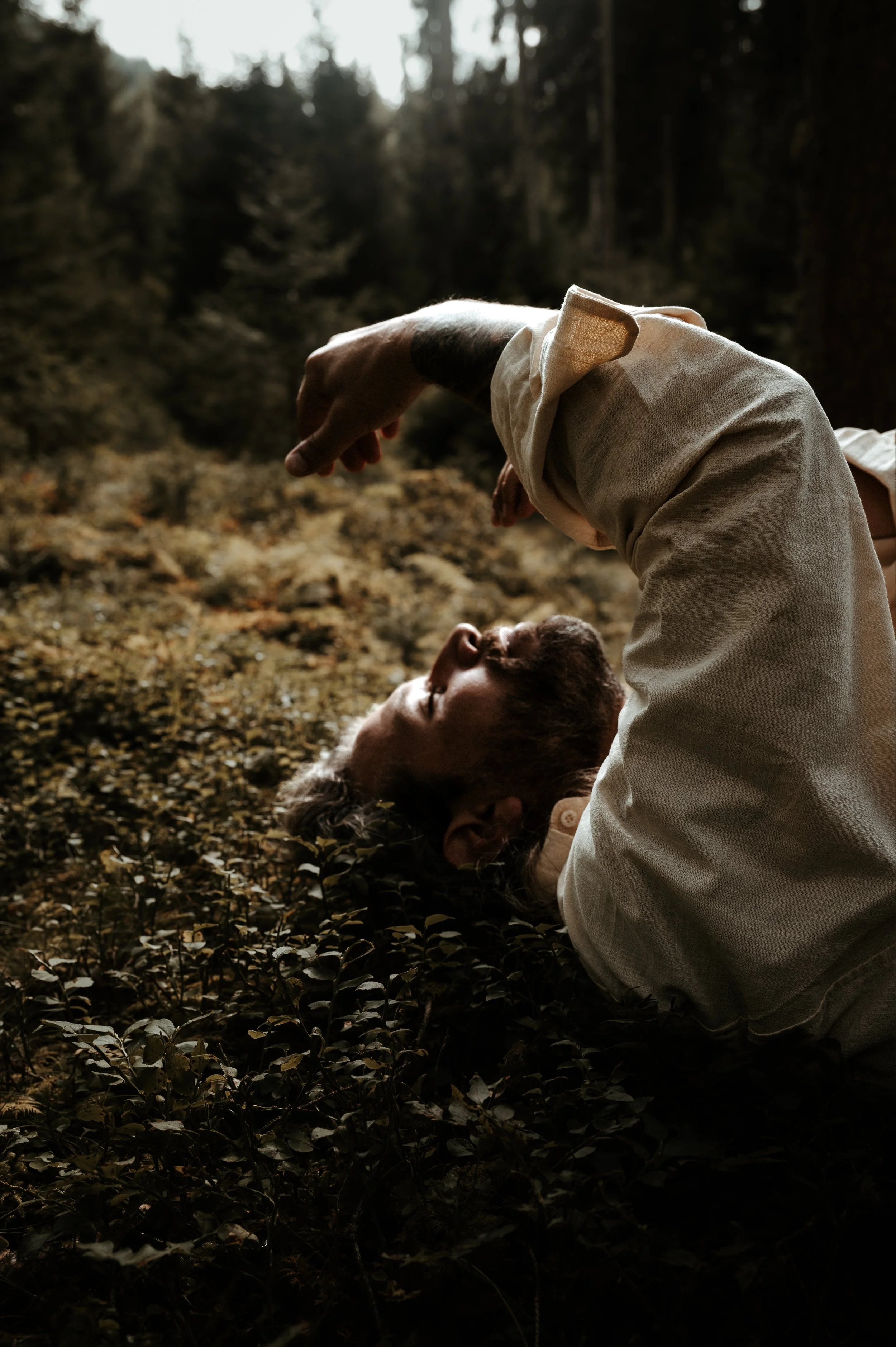 A person with reddish hair and tattoos on their arm lies on the ground in a forest, with another person in light-colored clothing leaning over them, surrounded by trees and fallen leaves.