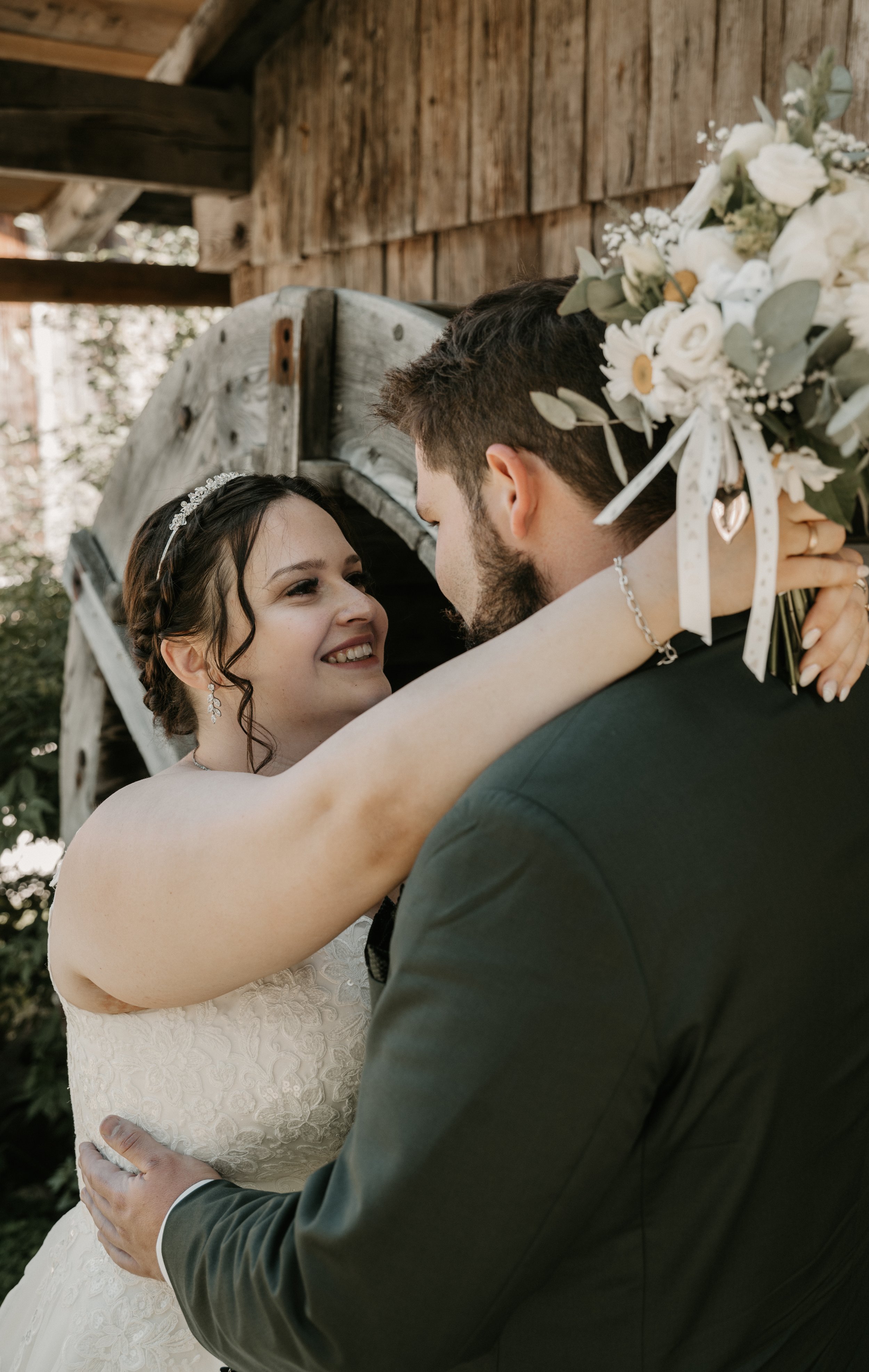 A bride and groom embrace outdoors, smiling at each other, with the bride holding a bouquet of white flowers and greenery, and a rustic wooden background.