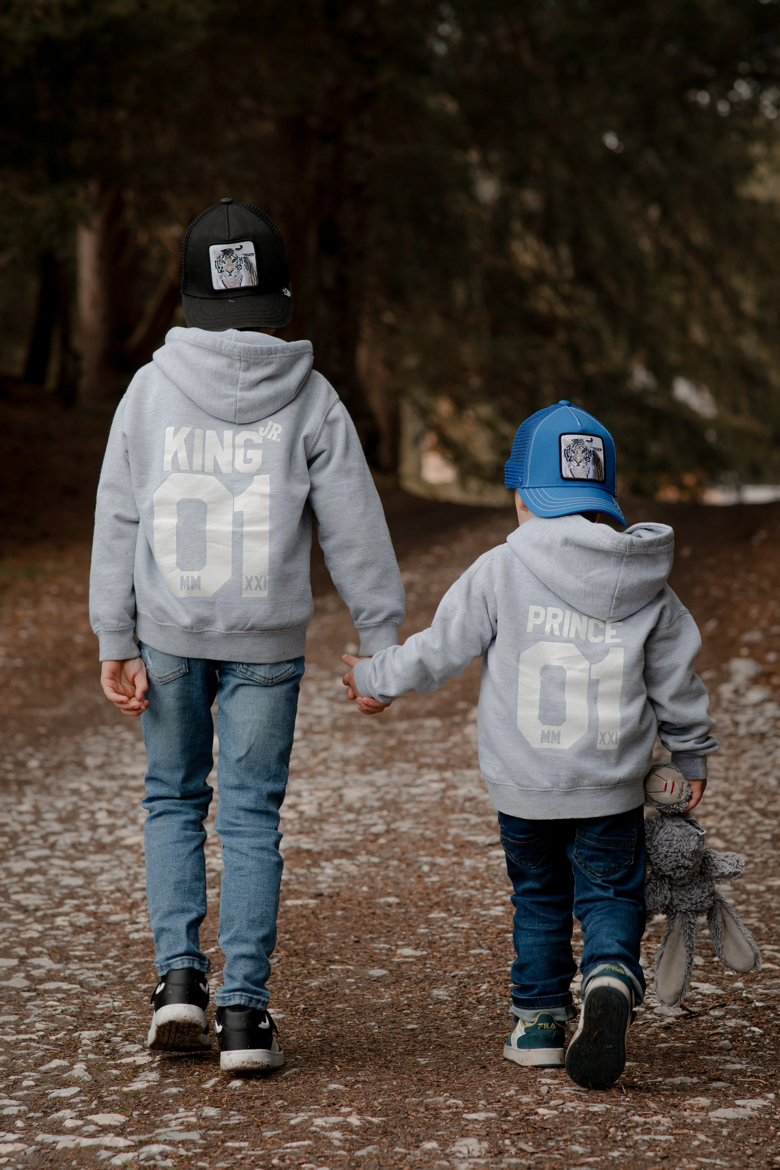 Two young boys walking hand-in-hand on a dirt and gravel park trail during autumn, dressed in grey hoodies with 'KING' and 'PRINCE' printed on the back, wearing baseball caps, one black with a tiger patch and the other blue with a similar tiger patch