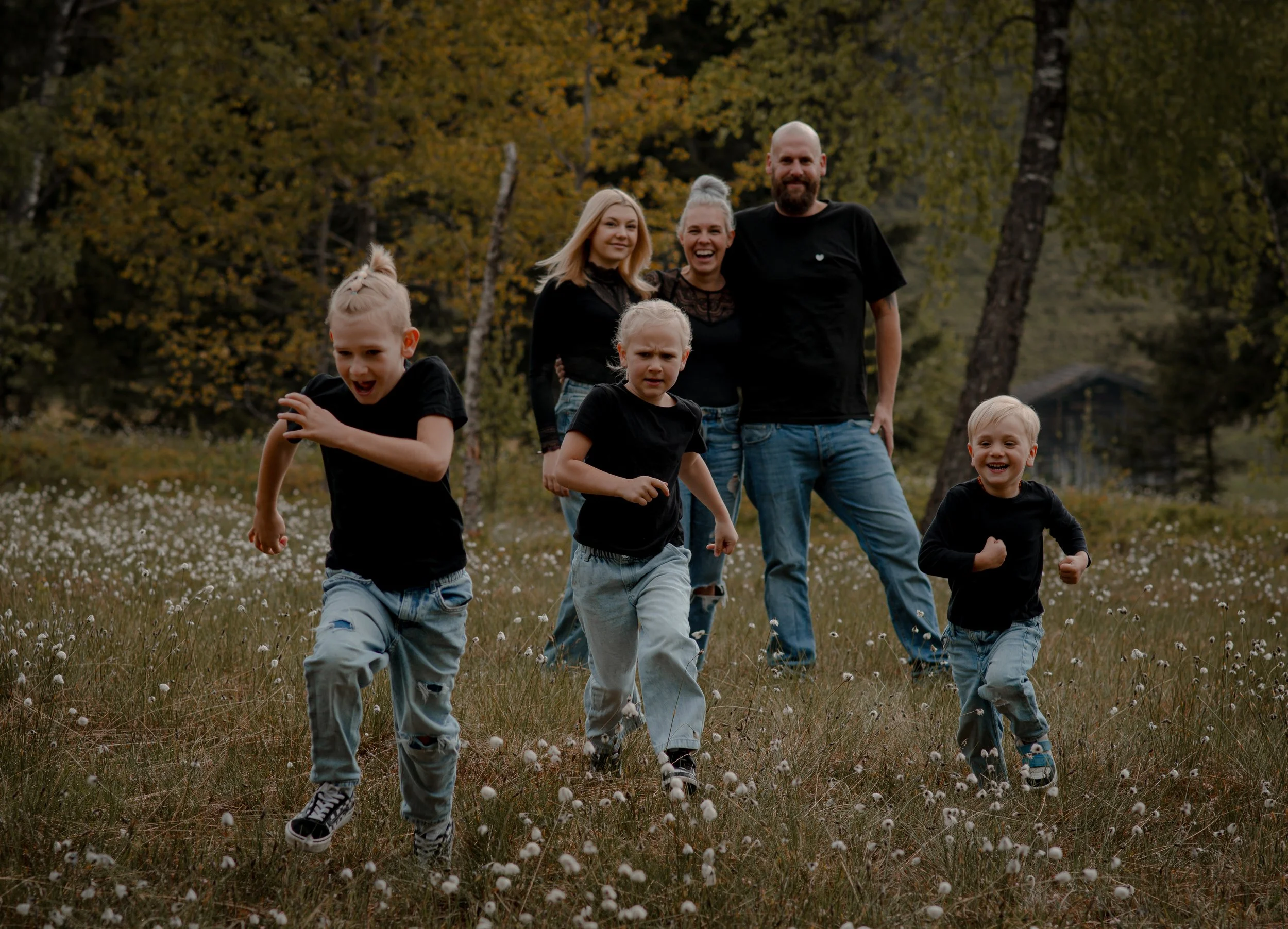 A family of six, including a man, a woman, and four children, walking and running outdoors in a grassy area during fall, with trees and a house in the background.