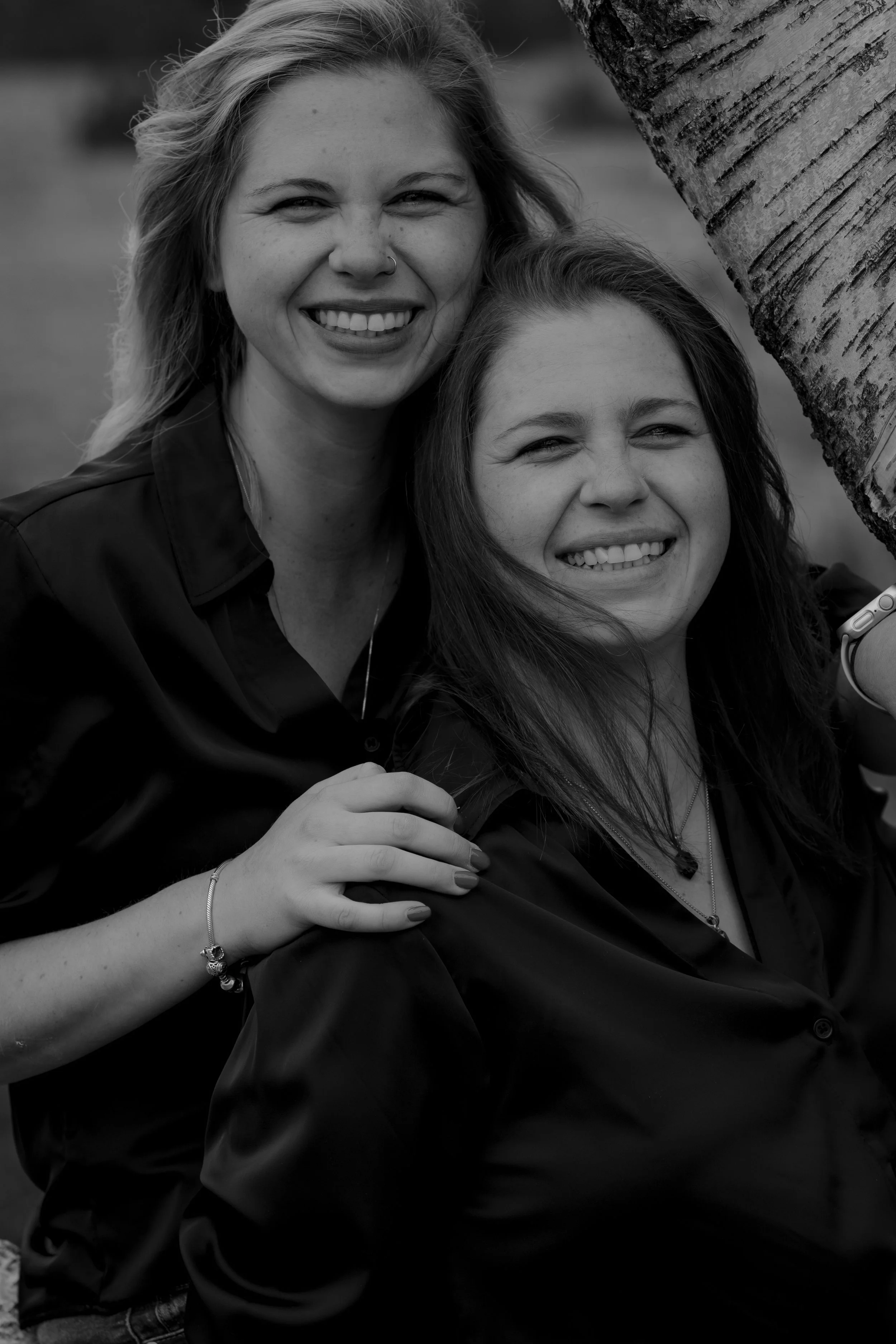 Two women smiling closely together outdoors, with one woman partially hidden behind a tree, in black and white photograph.