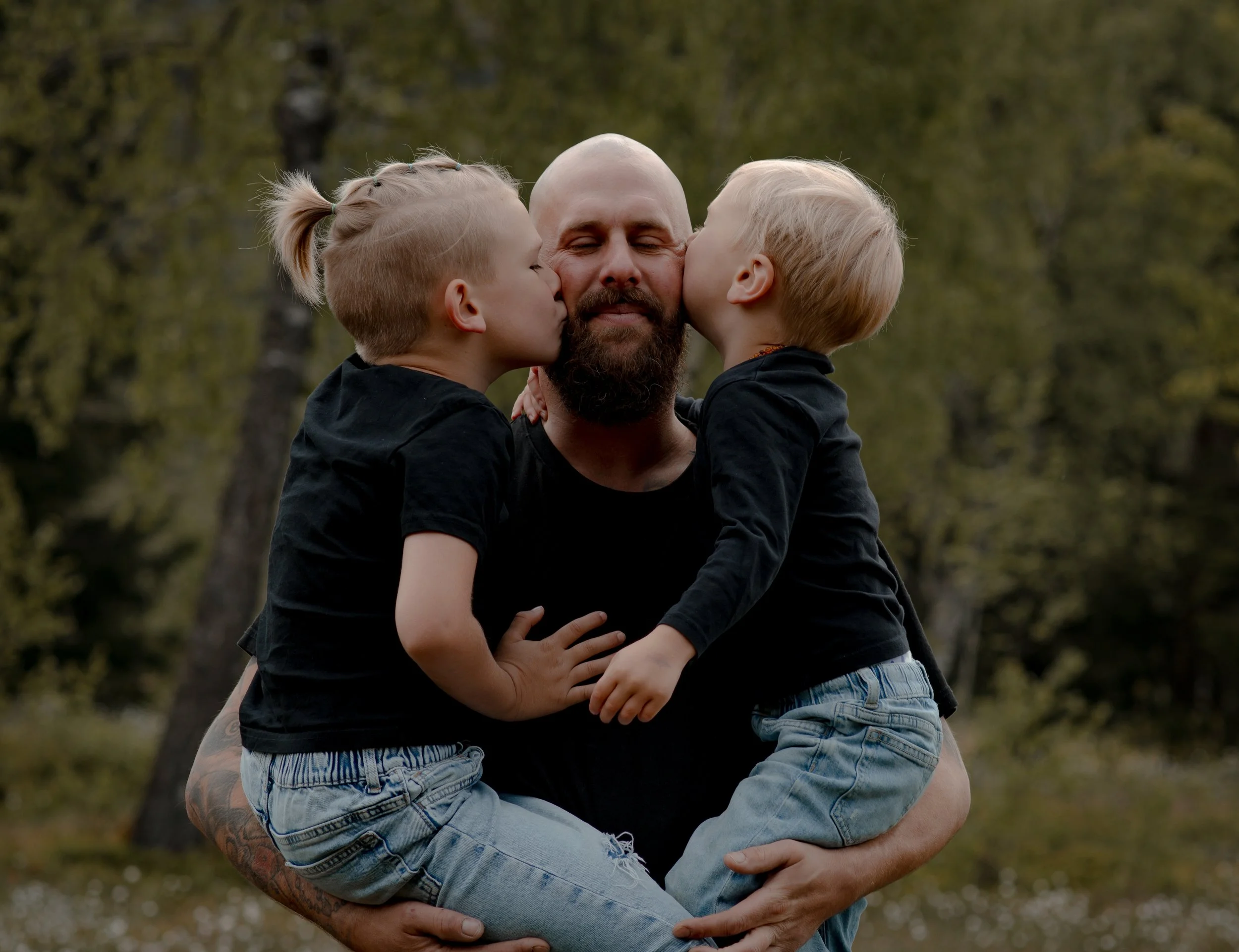 A father holding his two young children, giving each a kiss on the cheek outdoors in a park.