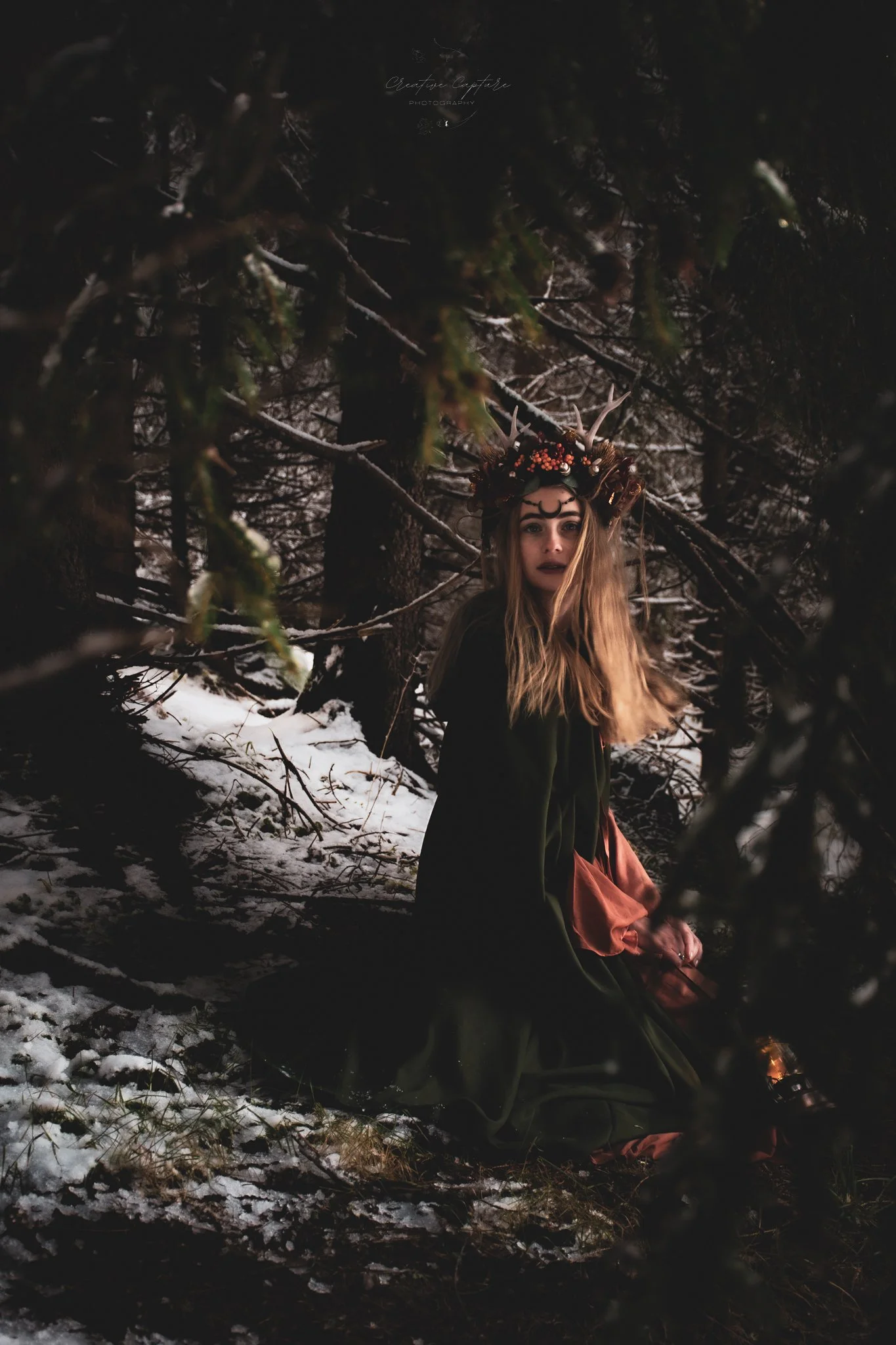 A woman with long hair wearing a crown of antlers and flowers, dressed in dark clothing with pink and green fabric, sitting in a snowy, dark forest at night, with a serious expression.