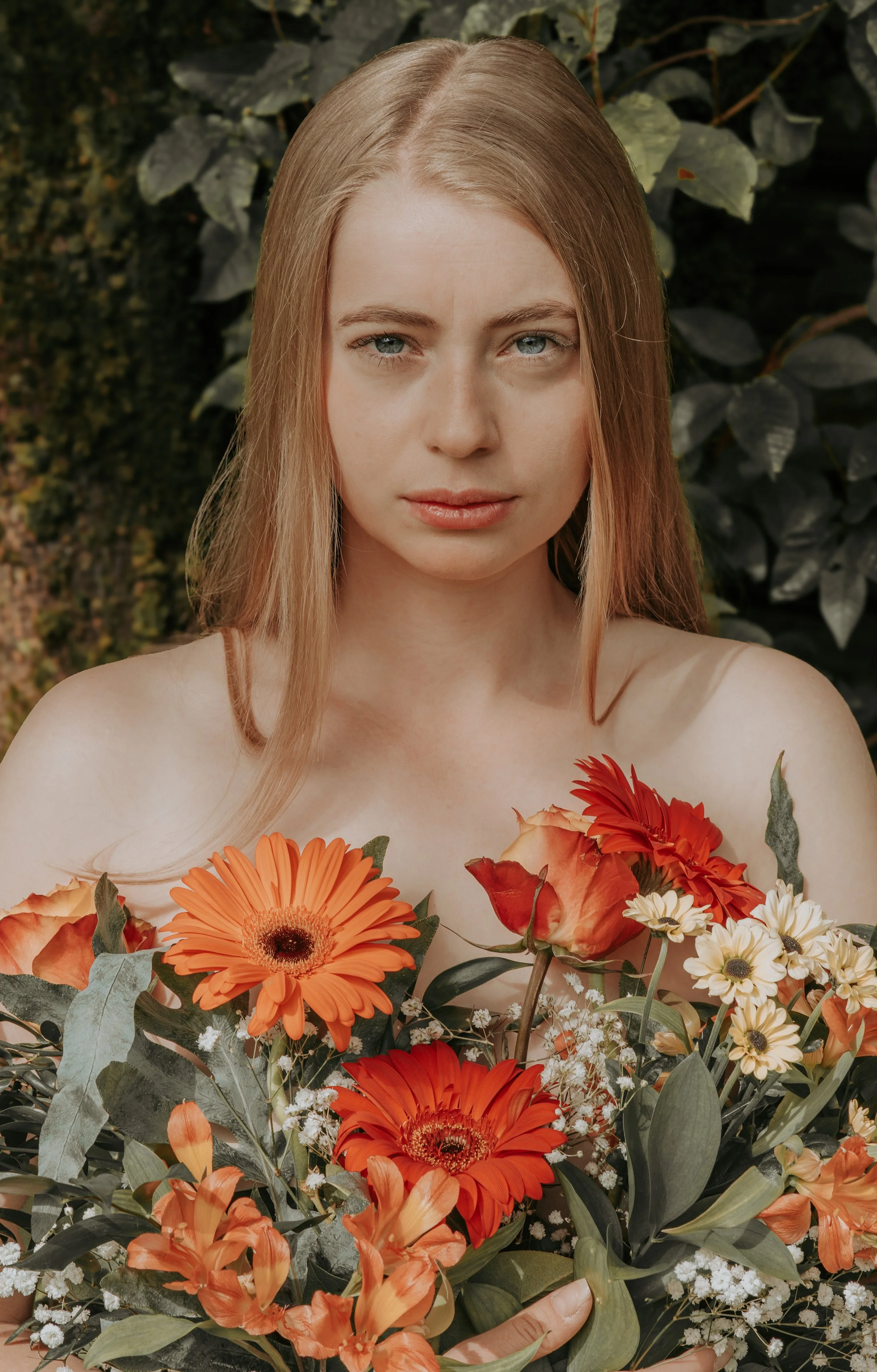 A woman with long blonde hair and blue eyes holding a large bouquet of orange, red, and white flowers against a dark leafy background.