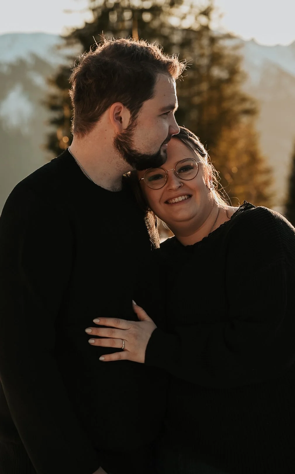 A couple standing outdoors during sunset, embracing and smiling. The man has a beard and short brown hair, while the woman wears glasses and has shoulder-length hair. They are dressed in black and are in a natural setting with trees and mountains in 