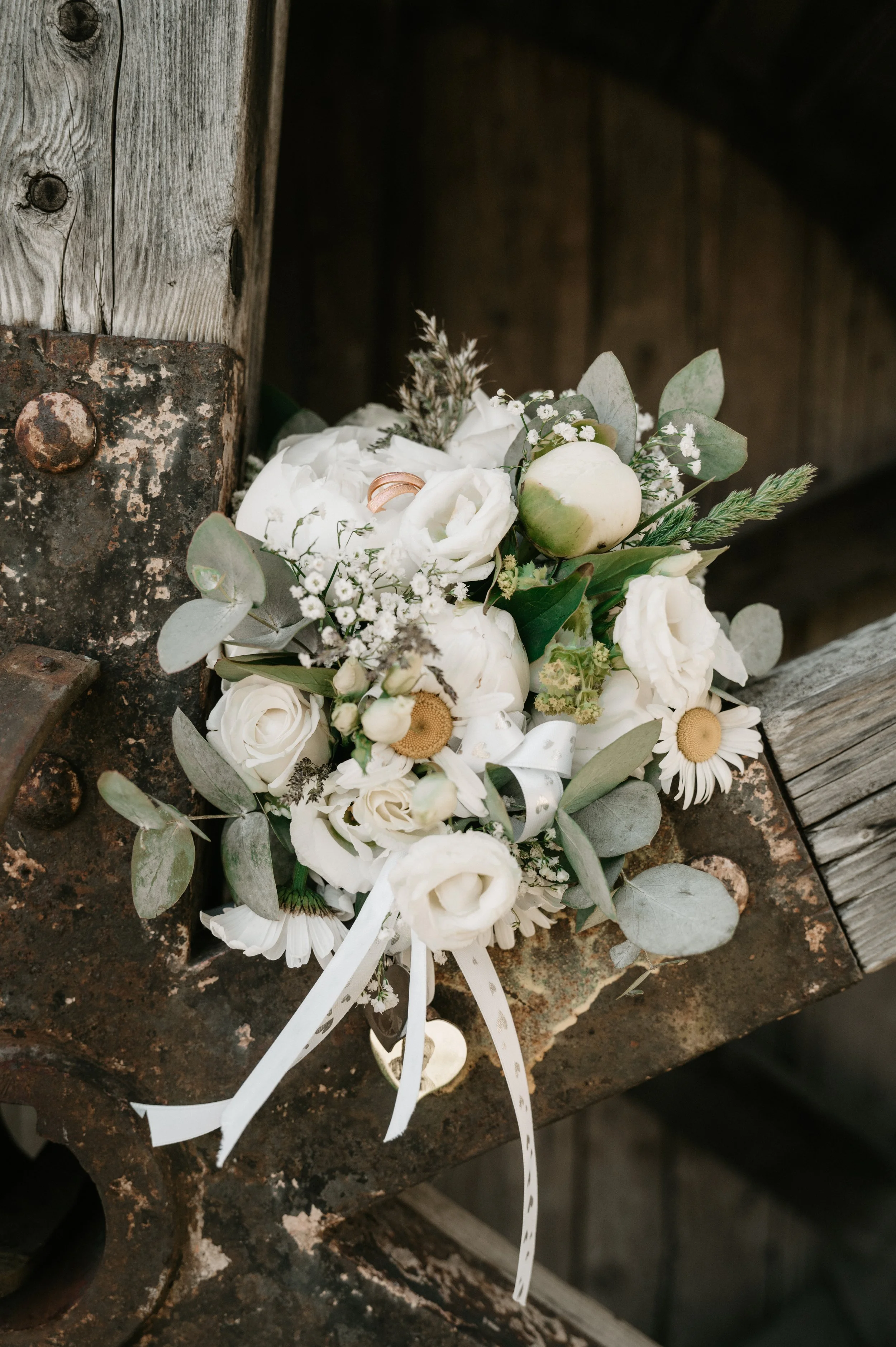 A bouquet of white flowers, including roses, daisies, baby's breath, and eucalyptus, tied with a white ribbon, resting on a rustic, rusty metal surface next to a weathered wooden wall.
