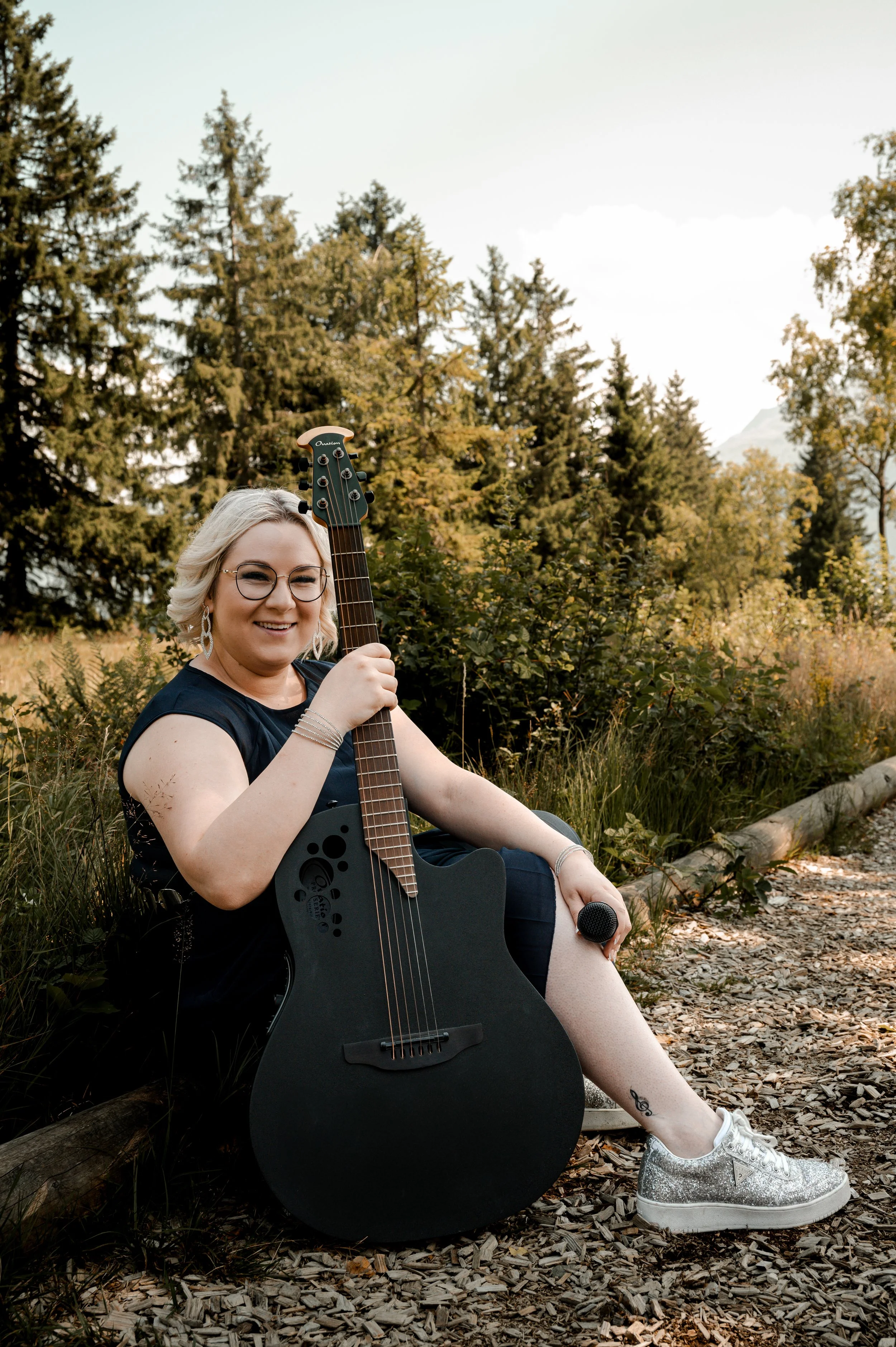 A woman with blonde hair and glasses sitting on the ground in a forest, holding an acoustic guitar and a microphone, smiling at the camera, wearing glittery sneakers and earrings.