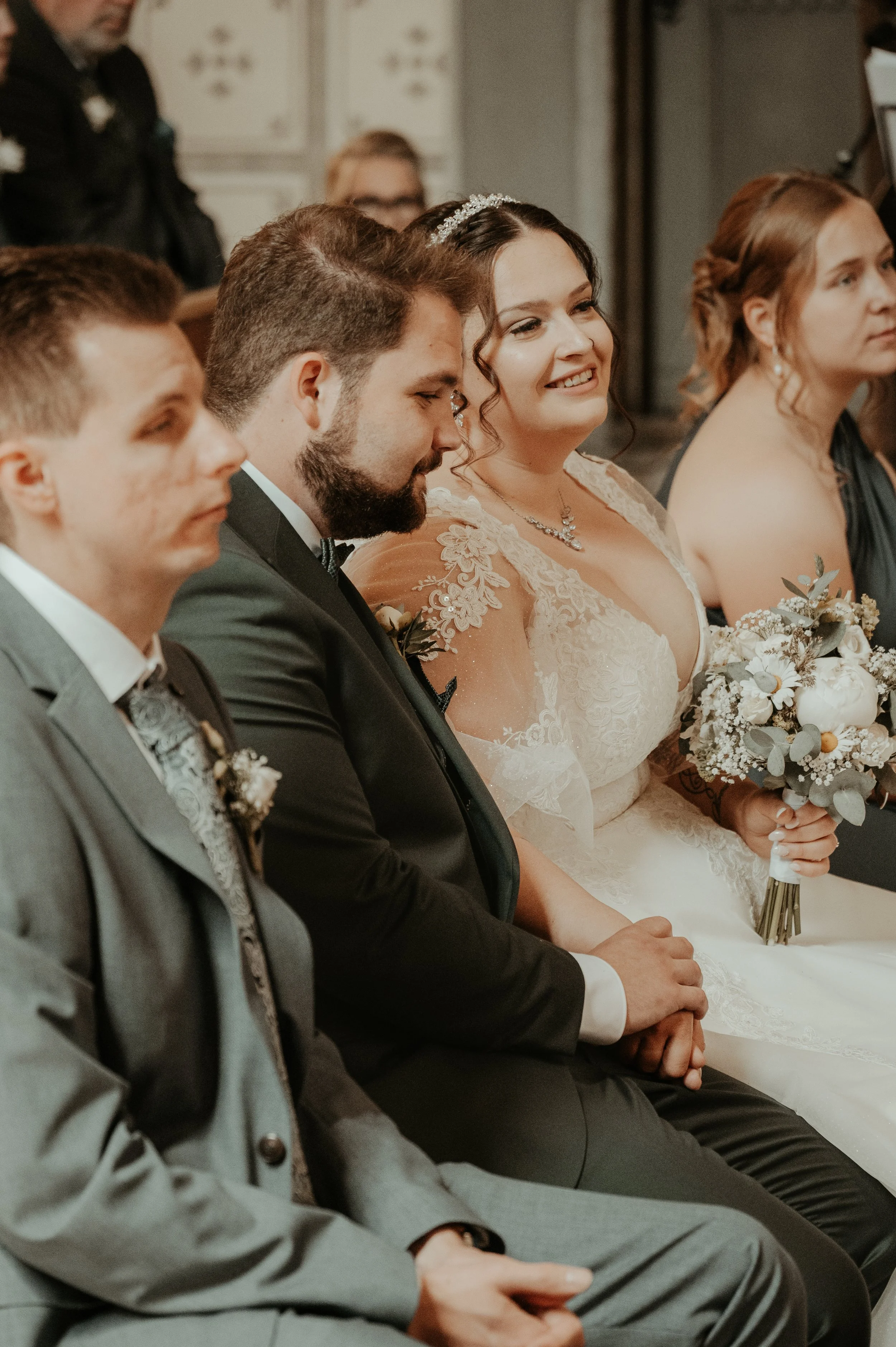 A group of people sitting in a row, dressed in formal attire, likely attending a wedding or ceremony. The bride, wearing a white lace gown and holding a bouquet, is smiling.