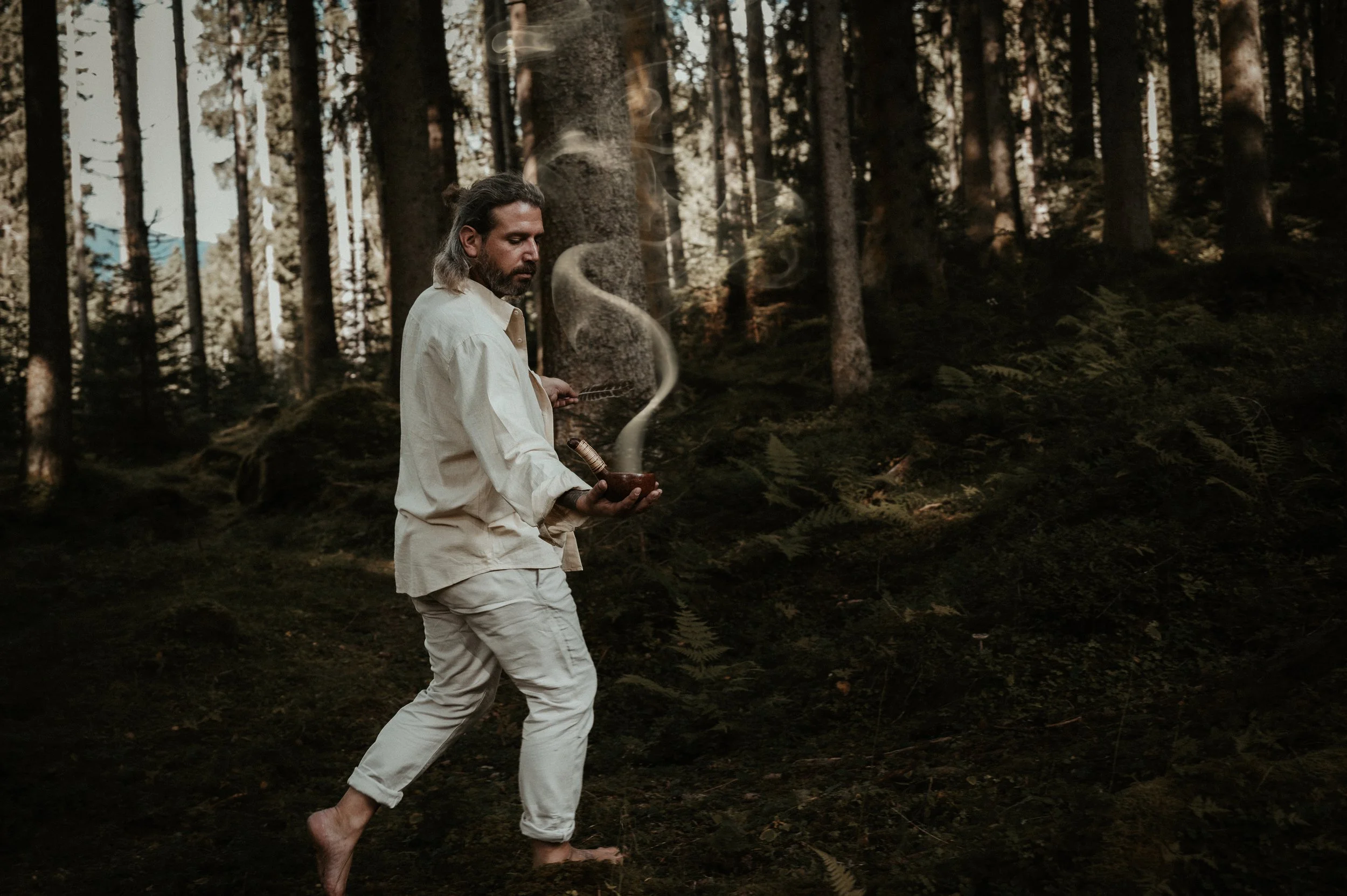 A man with long hair and a beard wearing a cream-colored outfit, barefoot, walking in a dense forest holding a smoking pipe.