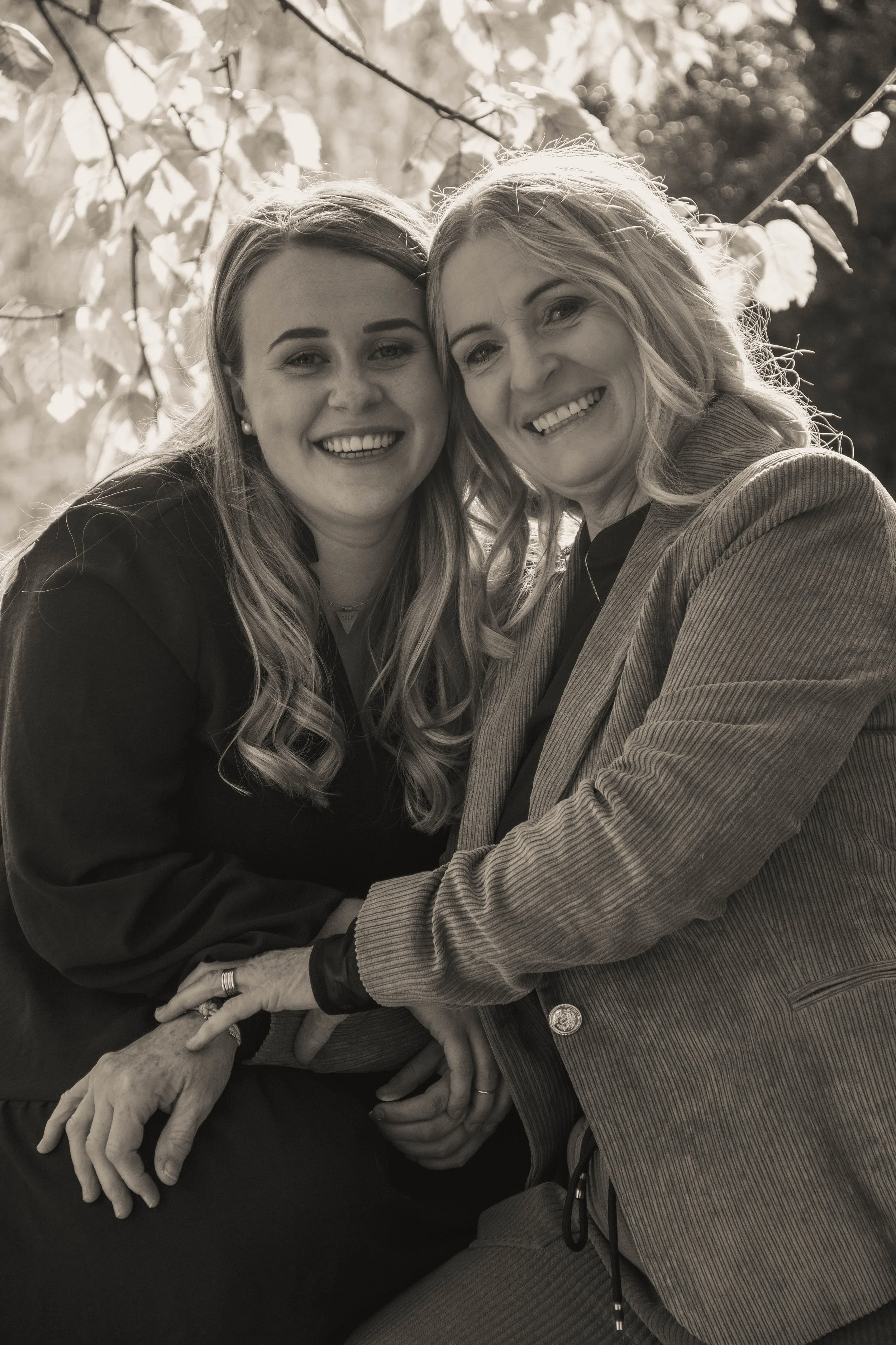 Two women smiling and holding hands in an outdoor setting with trees and leaves in the background.