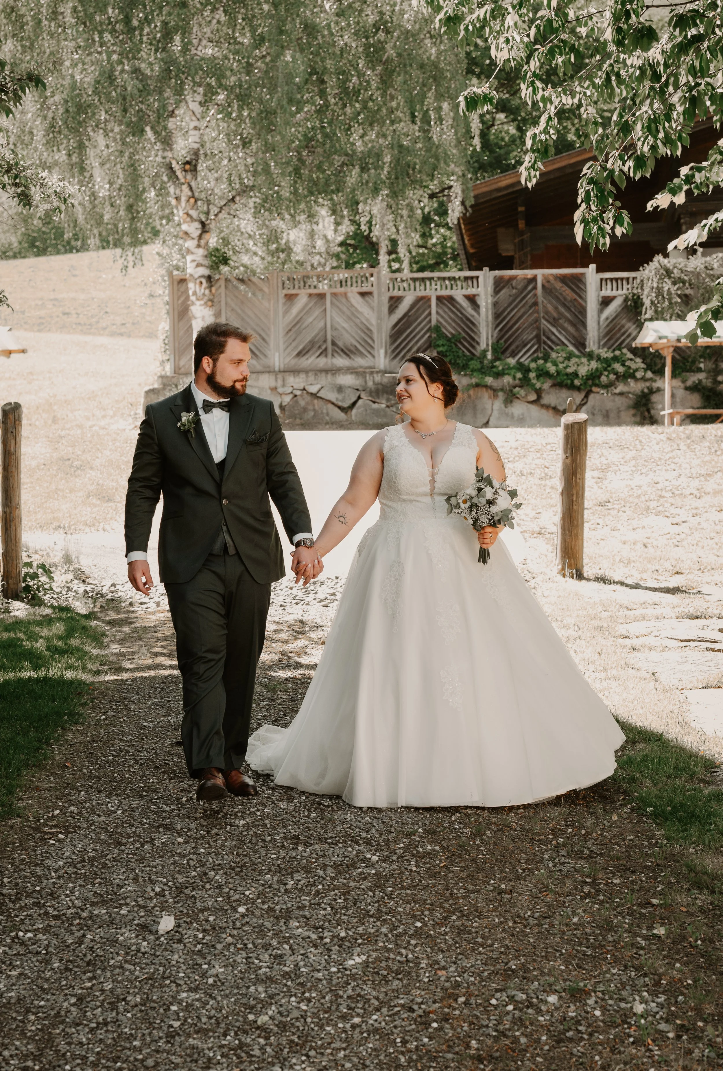 A bride and groom holding hands and walking outdoors in a garden or yard setting, smiling at each other. The bride wears a white wedding gown with lace details and a bouquet, and the groom wears a black suit with a bow tie.