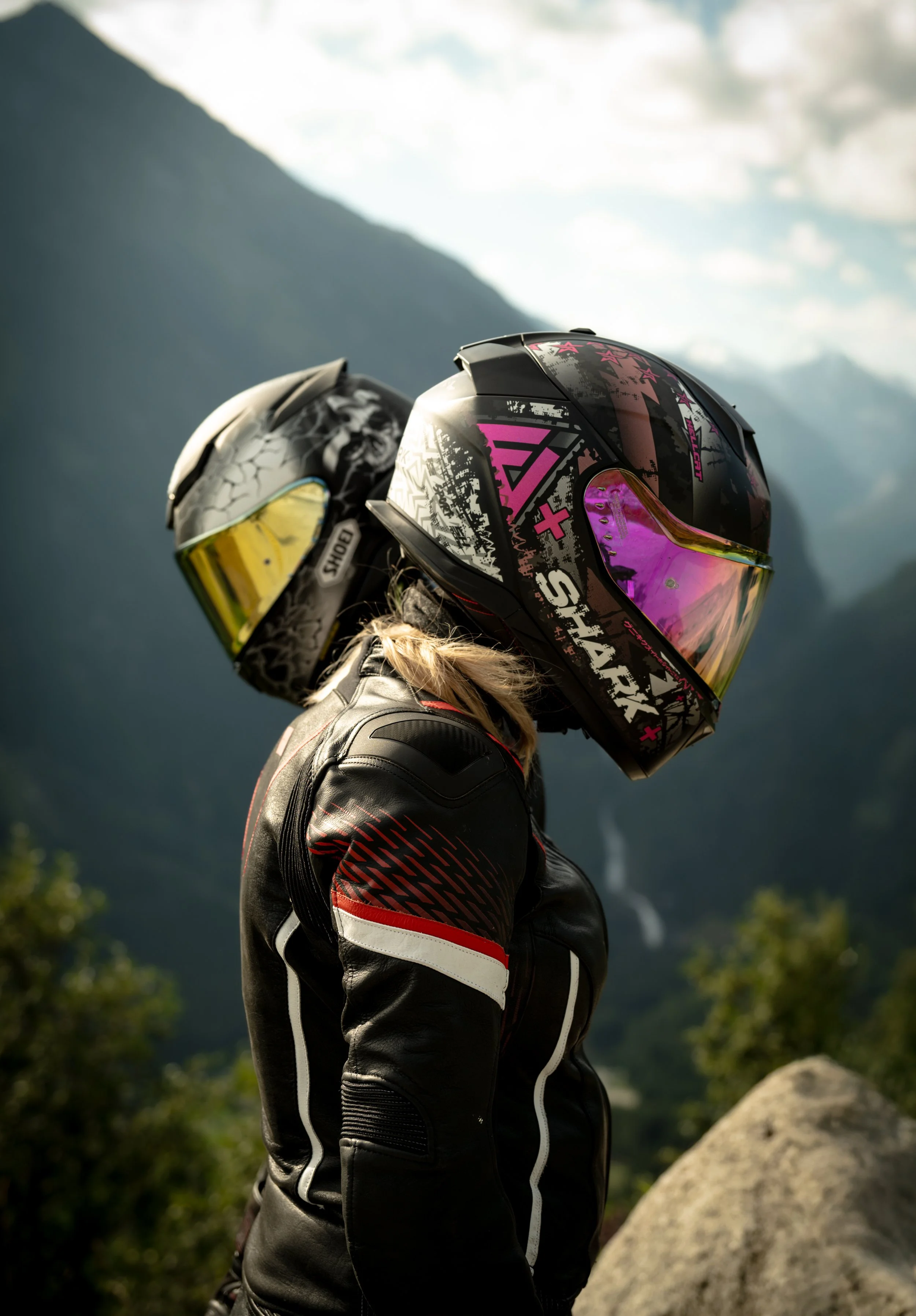 A person in motorcycle gear, including a helmet with pink, black, and yellow reflective visor, standing outdoors with mountains and trees in the background.