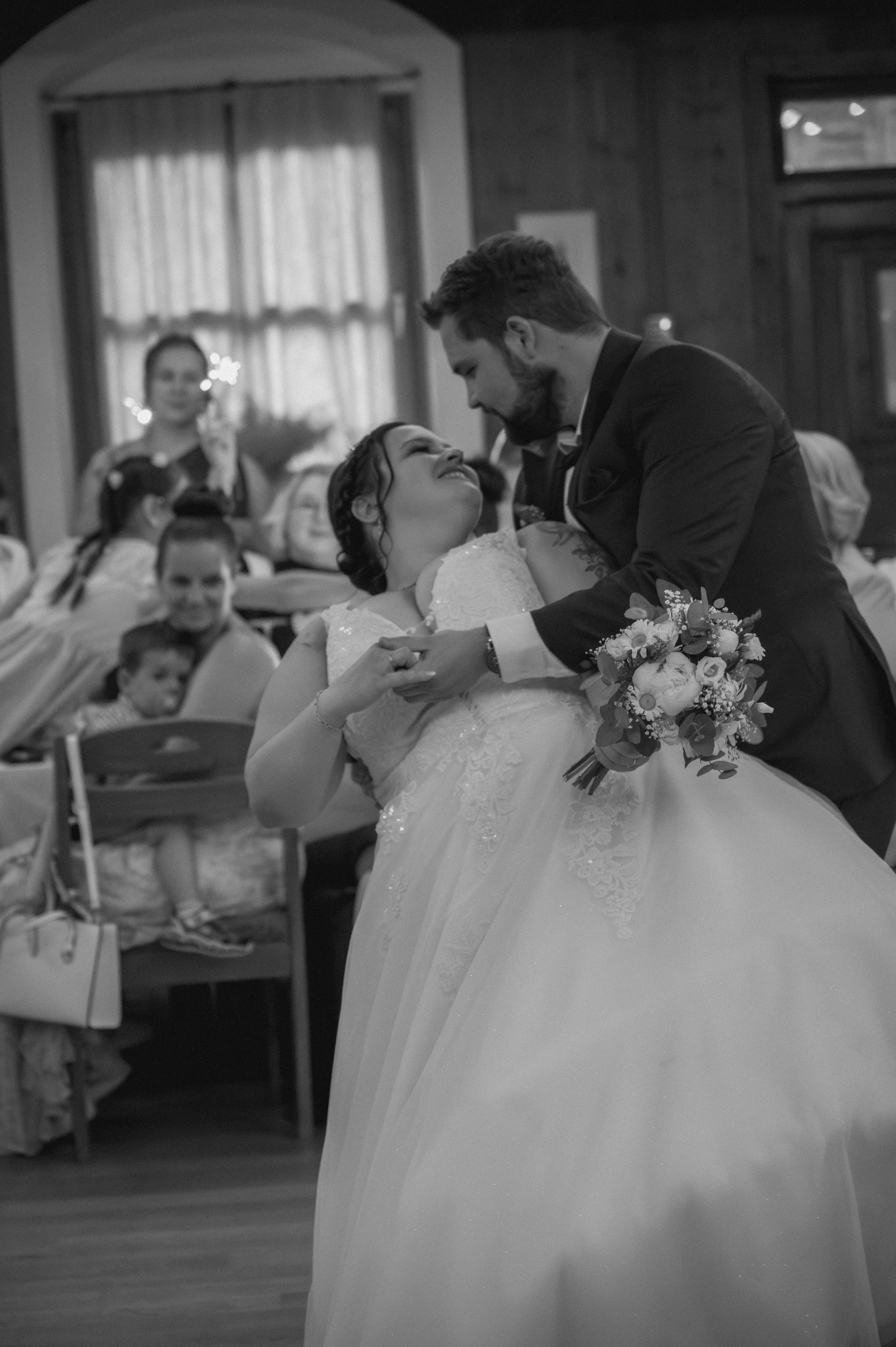 A black and white photo of a wedding scene, with a bride and groom dancing, surrounded by seated guests and children, in a room with curtains and wooden walls.