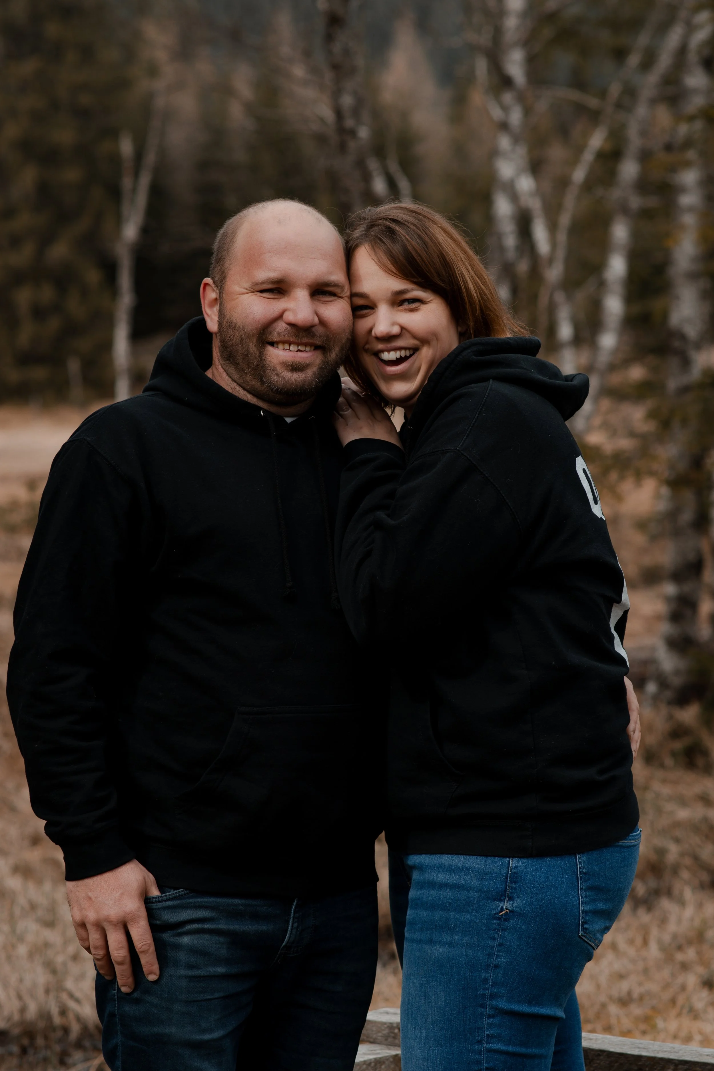 A couple stands closely together outdoors, smiling and laughing, with trees in the background.