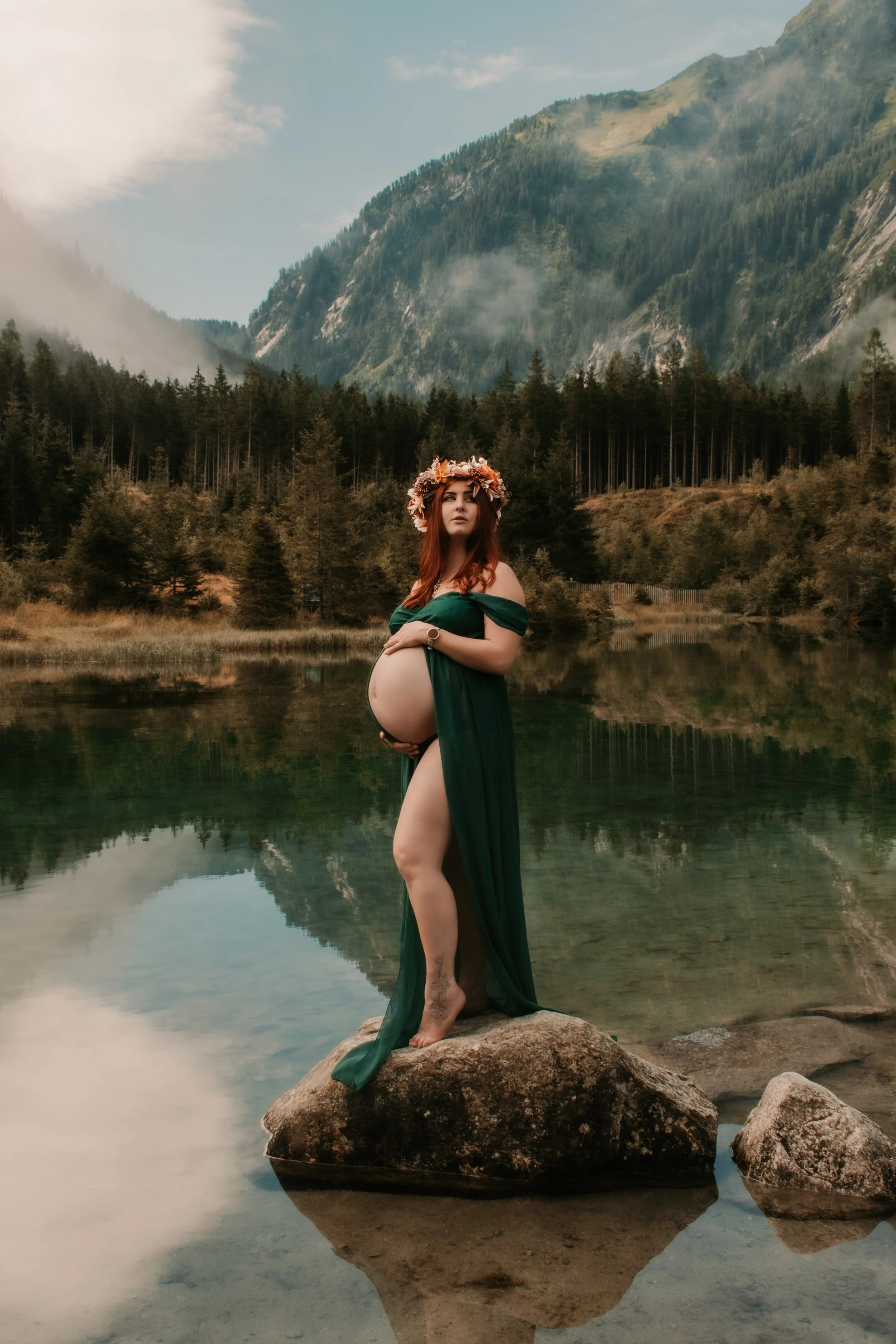 A pregnant woman wearing a green dress and a flower crown, standing on a large rock in a lake, with a mountain and forest scenery in the background.