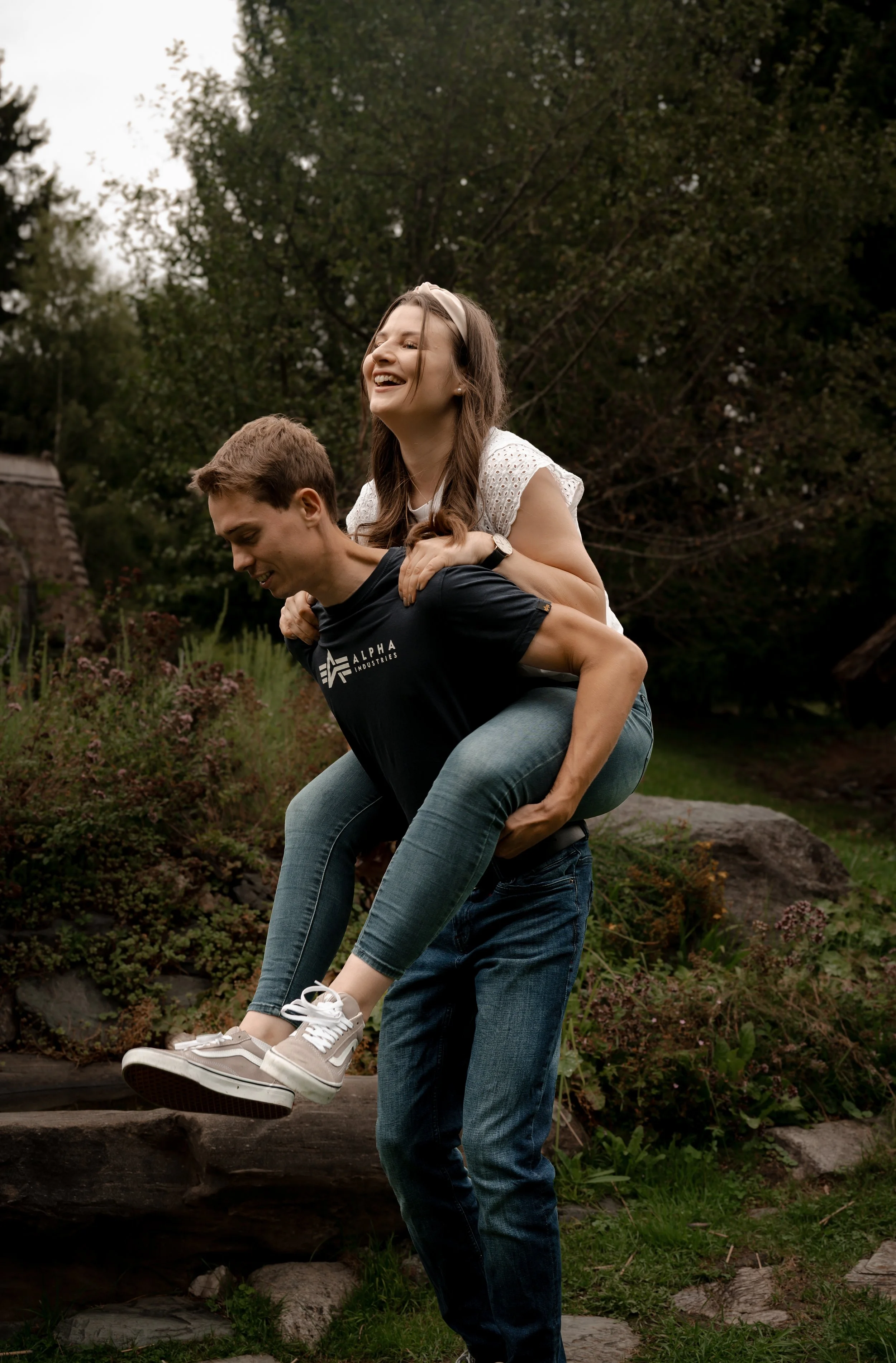 A young woman with long brown hair smiling while sitting on the shoulders of a young man with short brown hair in a garden with trees and plants.