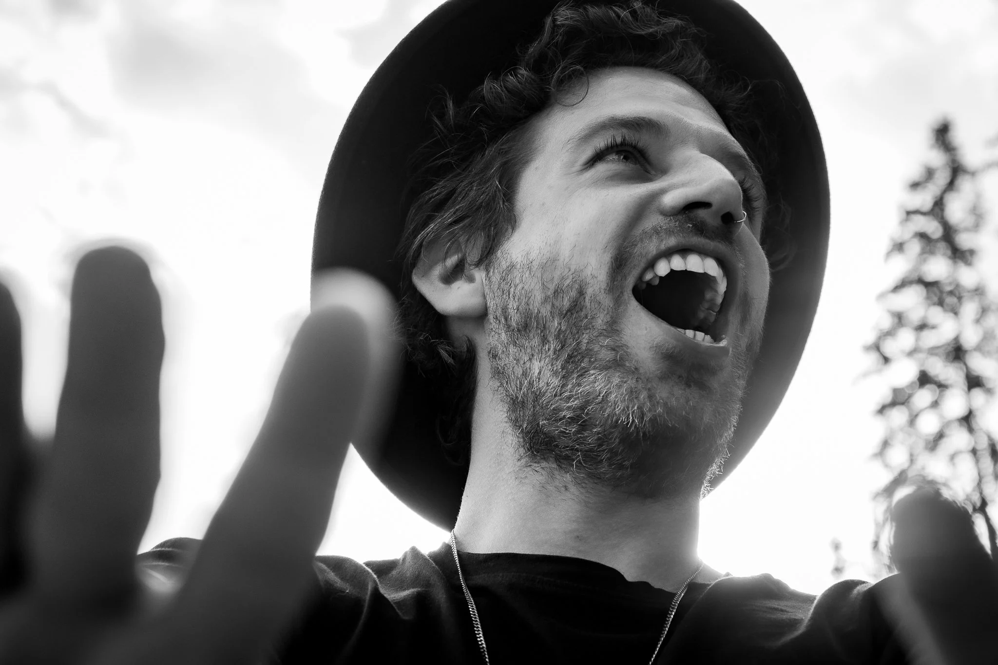 Black-and-white photo of a young man with a beard and curly hair, wearing a wide-brimmed hat, smiling and looking upward, with some fingers partially visible in the foreground.