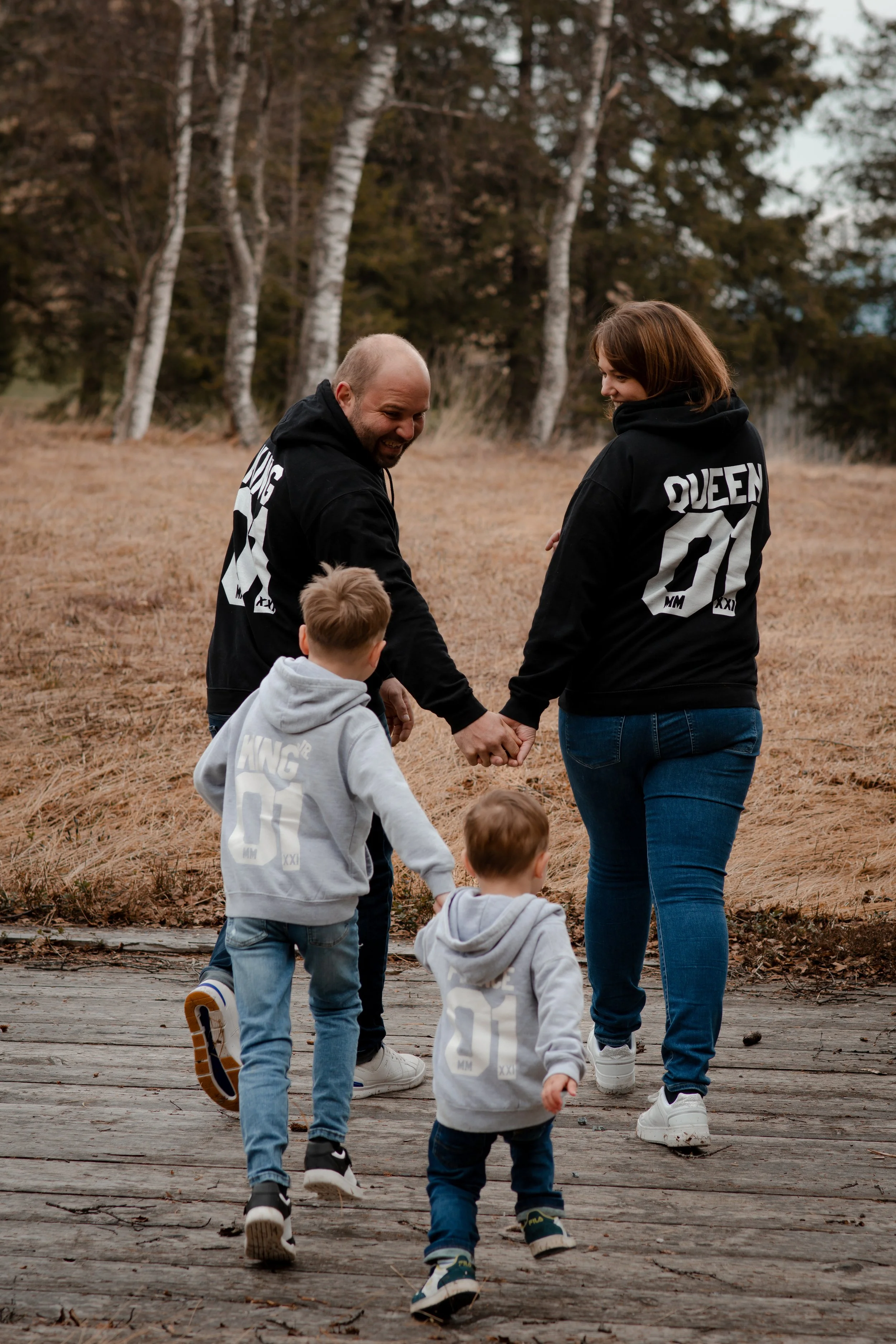 A family holding hands and walking outdoors on a wooden path in a park with tall trees and dry grass, enjoying a day out together.