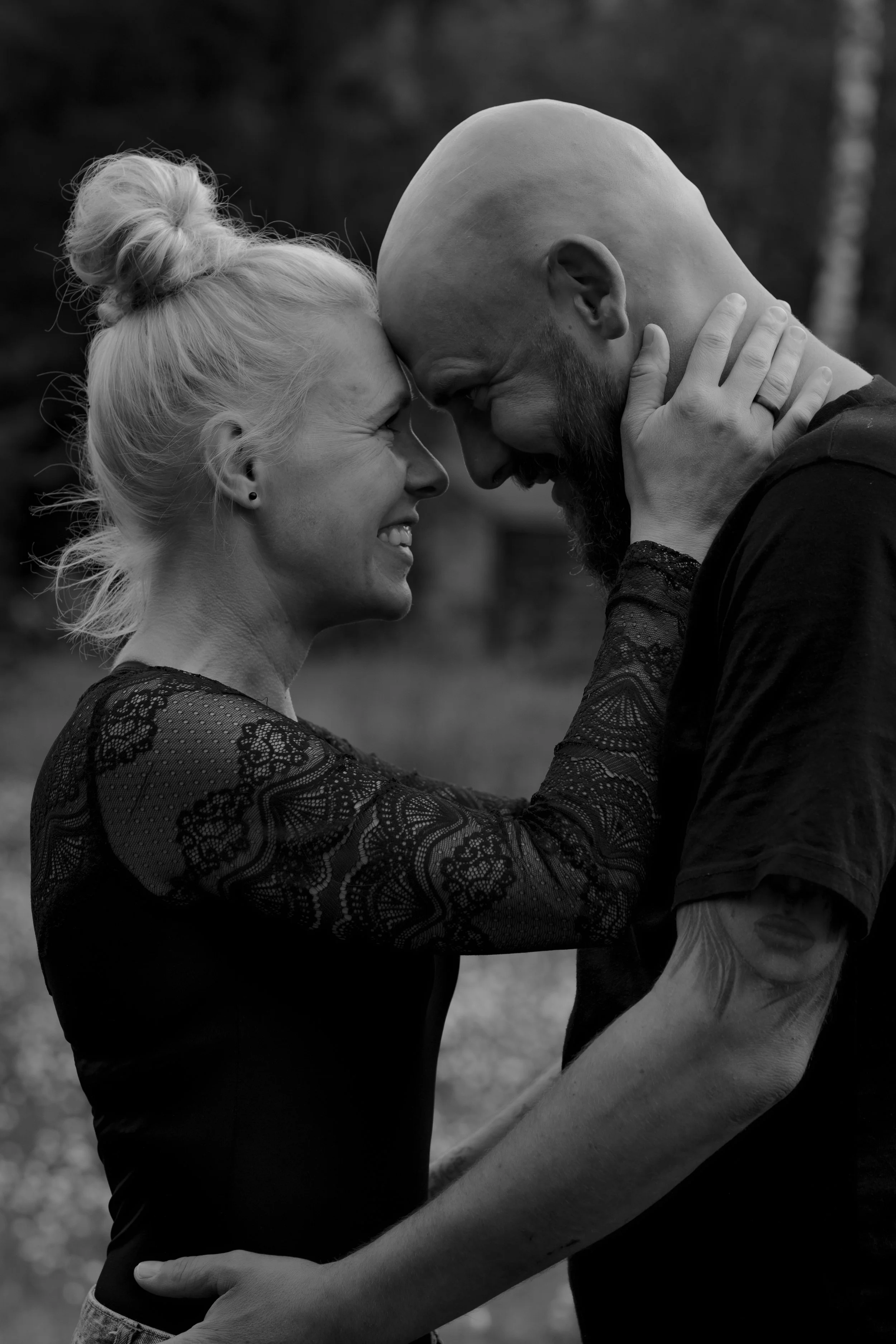 A black and white photo of a couple touching foreheads and smiling, with their arms around each other, outdoors in a natural setting.