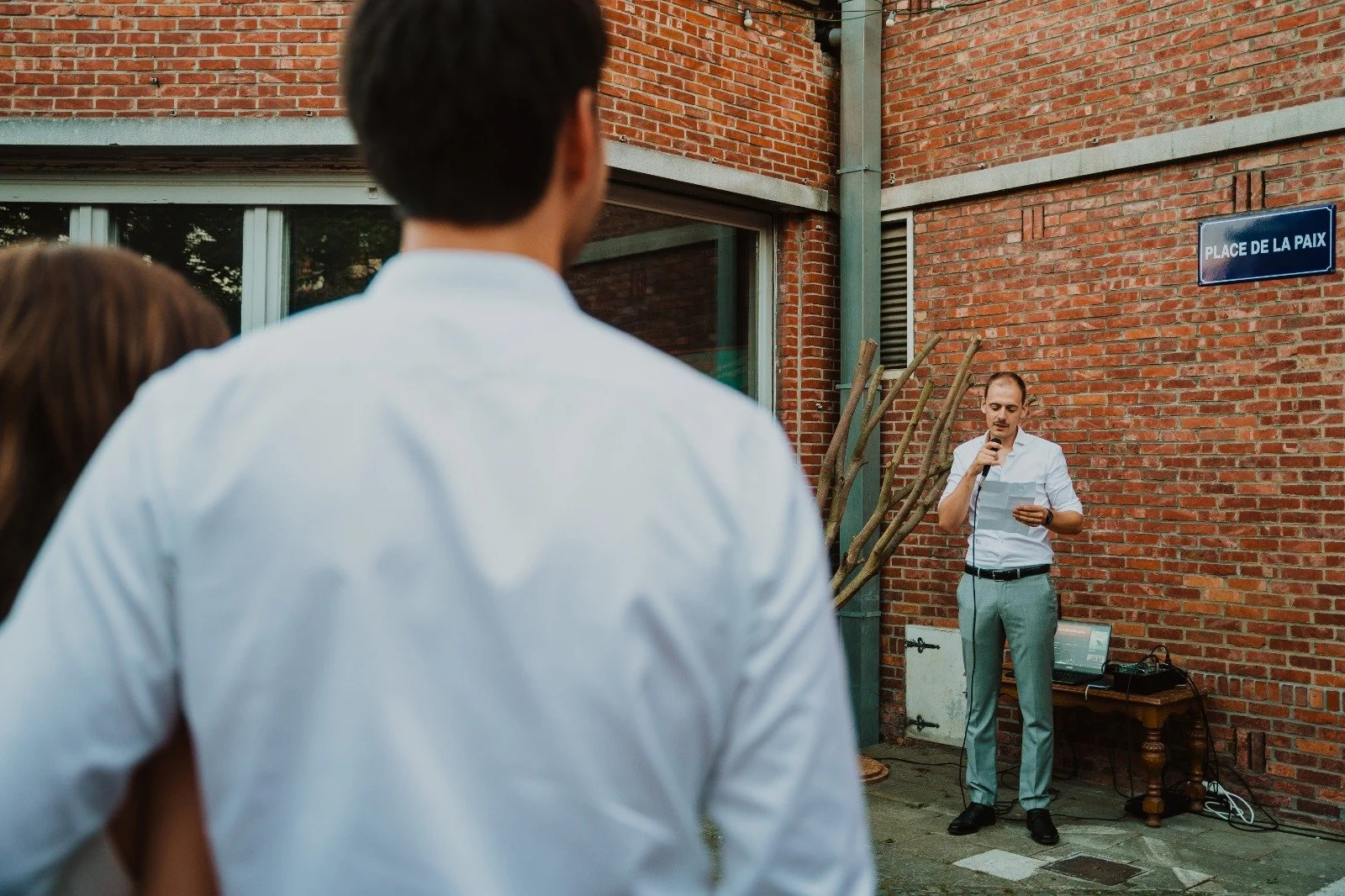 A man in a white shirt is giving a speech or presentation outdoors, holding a microphone and reading from papers, against a brick wall with a street sign that says 'Place de la Paix'. Two people are in the foreground, watching him.