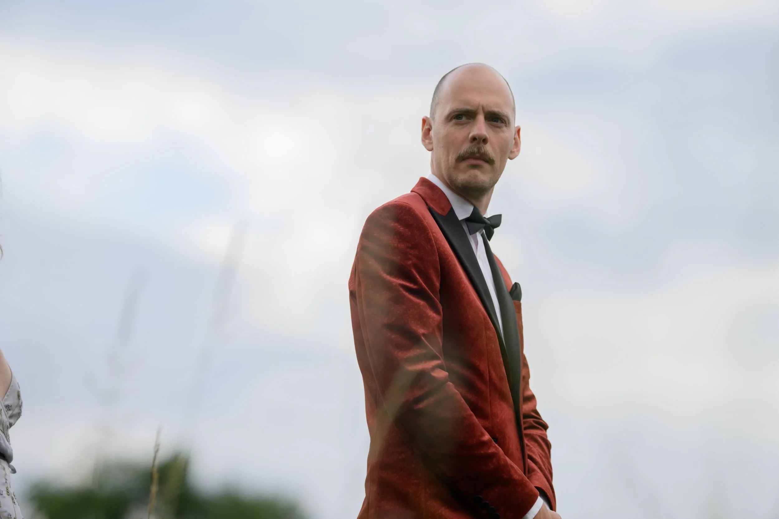 A man with a bald head and mustache wearing a red velvet tuxedo jacket, white shirt, and black bow tie, standing outdoors against a cloudy sky.