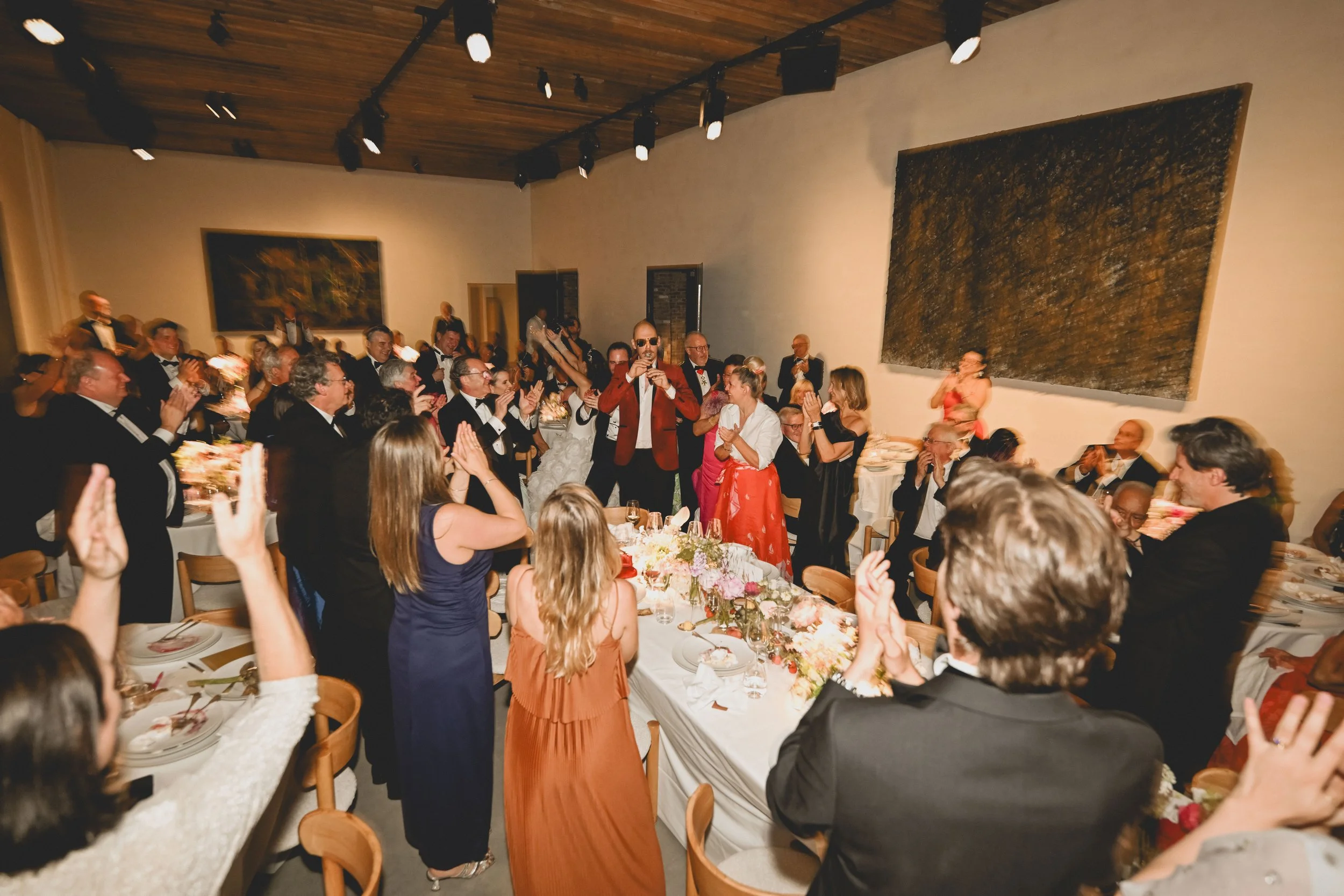 A large group of guests dressed in formal attire attending a celebration or banquet in a decorated hall, some clapping and others standing while a man in a red suit sings or speaks into a microphone.