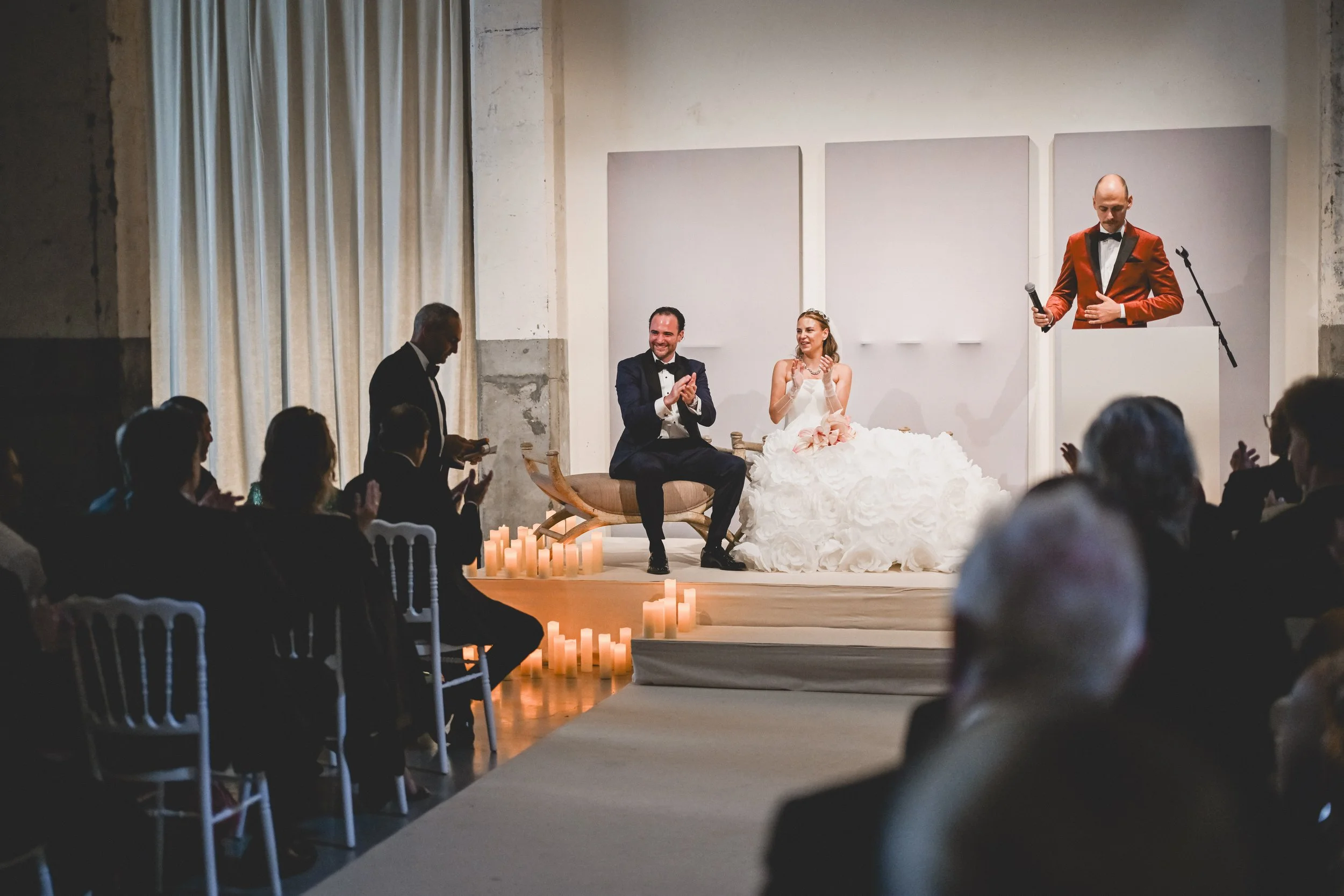 A wedding ceremony with a bride and groom seated on stage, a person at a podium, and guests clapping, candles on the floor.