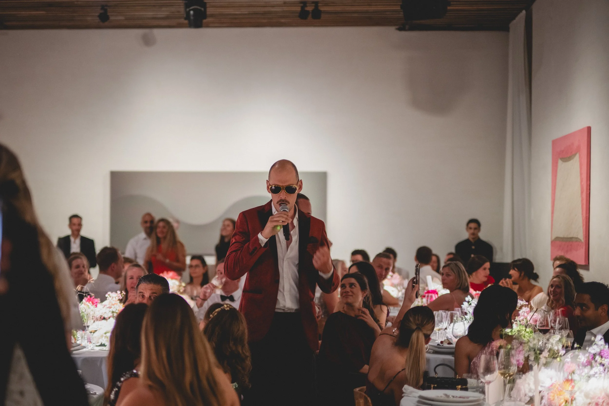 A man wearing sunglasses and a red suit jacket is speaking into a microphone at a formal event. He is surrounded by seated guests at decorated tables with pink and white floral centerpieces, in an art gallery or banquet hall with white walls and artwork hanging.