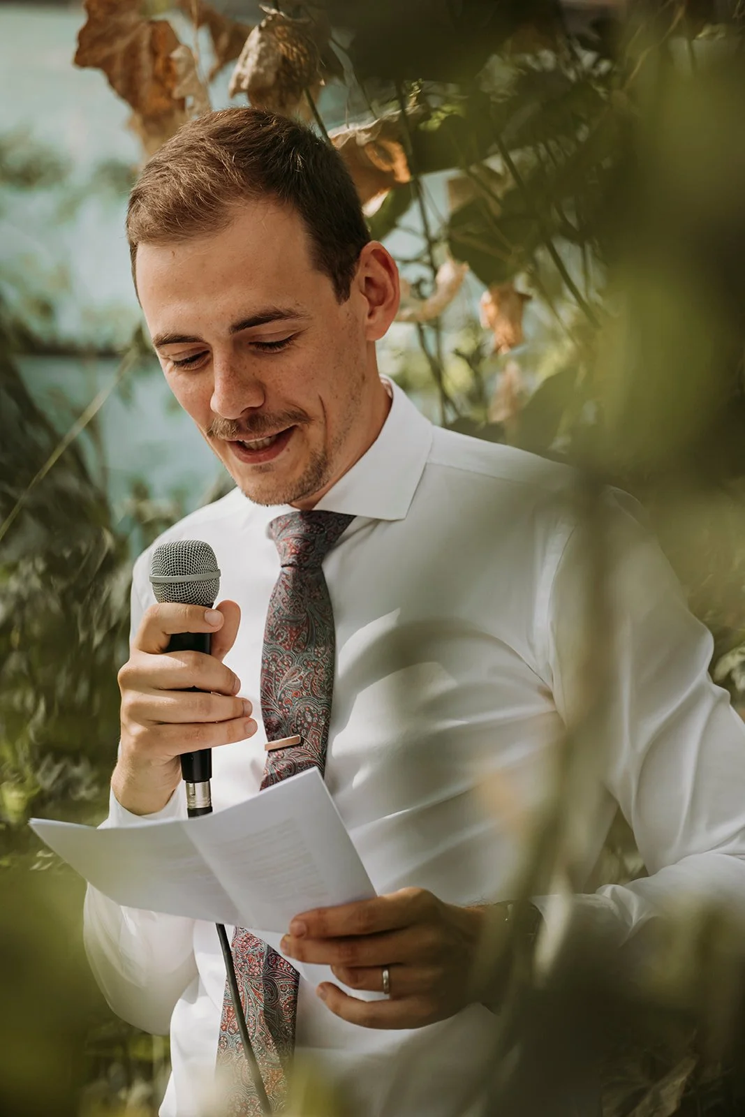 A man in a white shirt and patterned tie speaking into a microphone, reading from a piece of paper, surrounded by greenery.