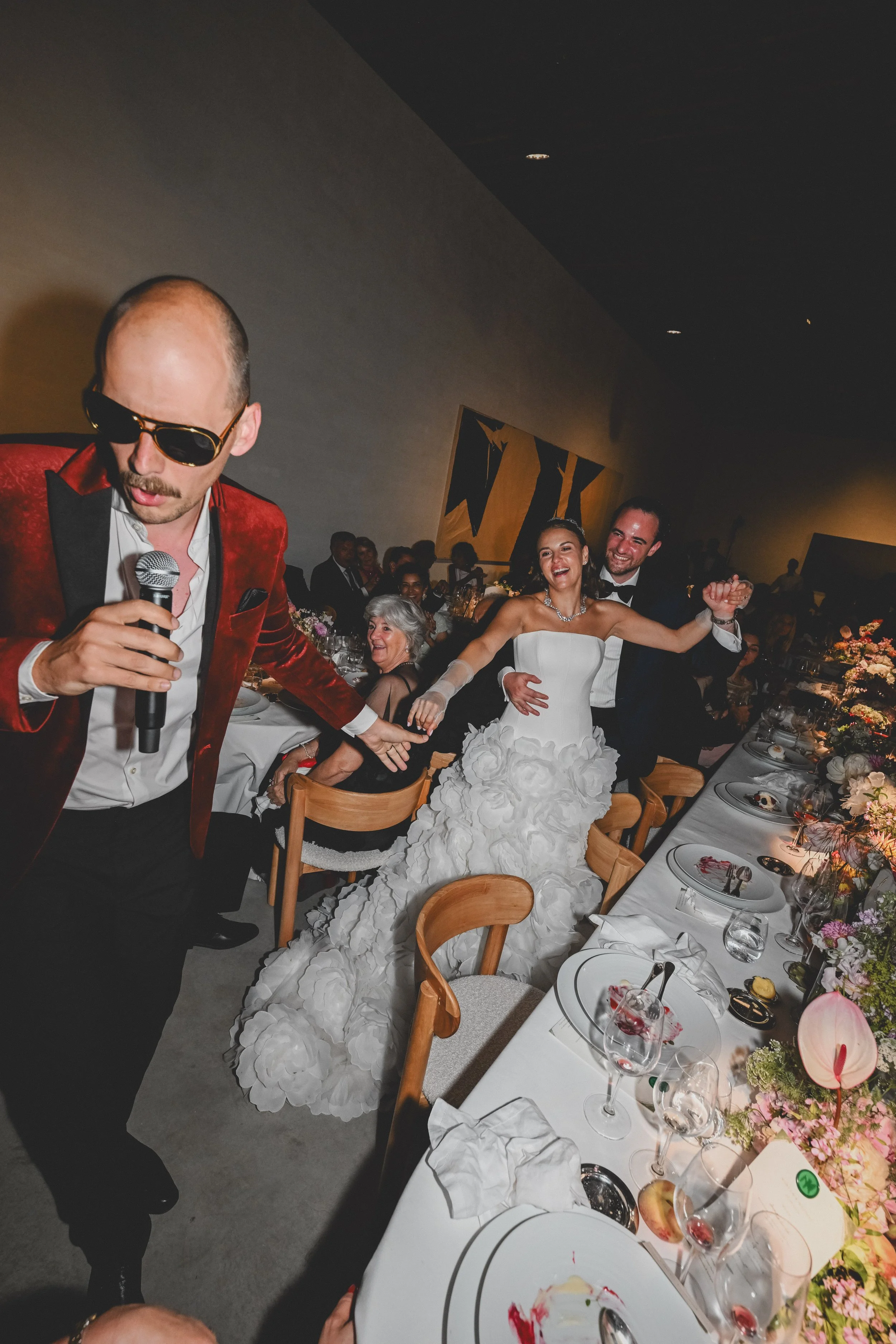 A wedding reception with a bride and groom dancing, surrounded by guests seated at decorated tables, with a man in a red blazer and sunglasses holding a microphone in the foreground.