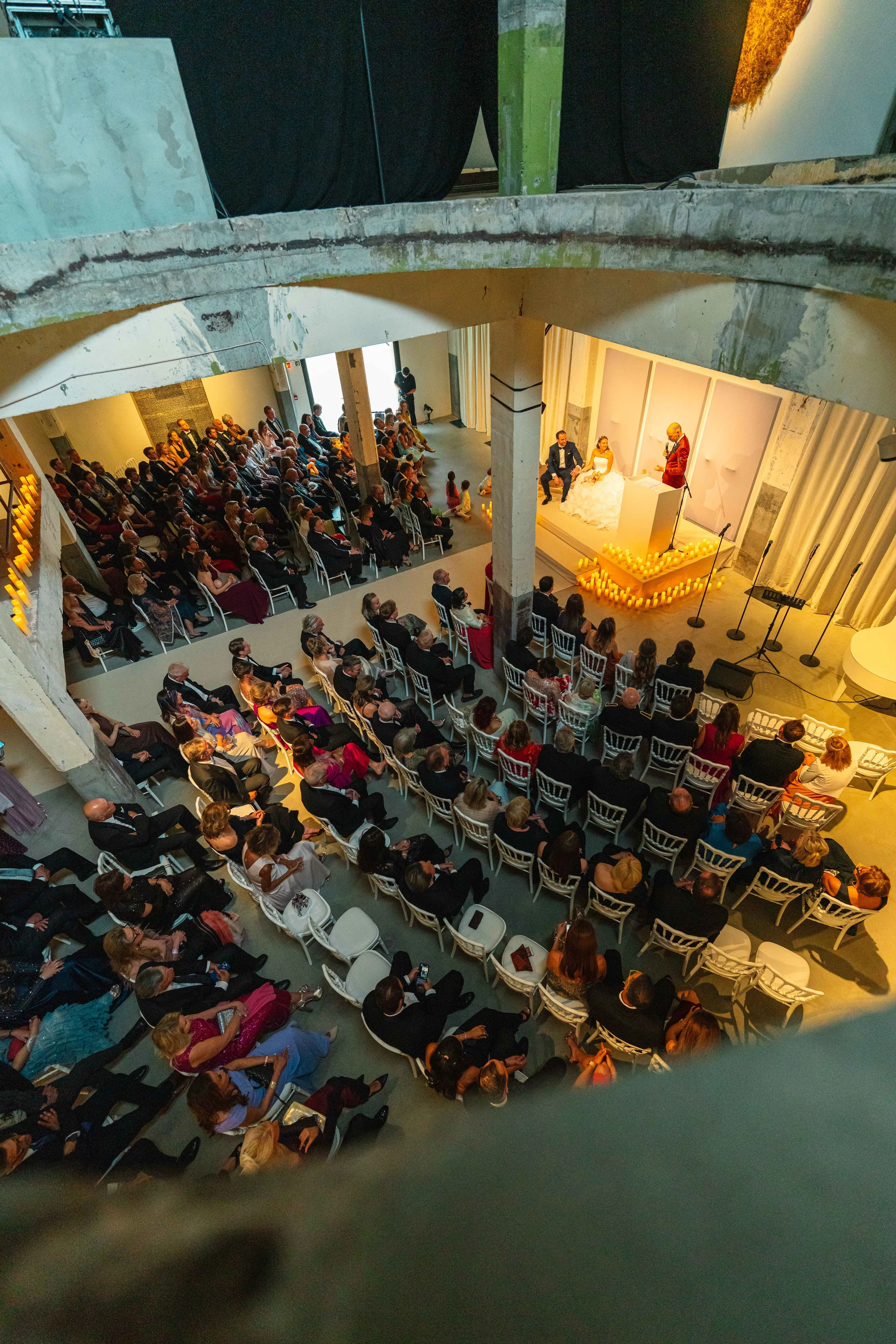 A large gathering of people seated in rows facing a stage where a wedding ceremony is taking place, with a bride and groom sitting and a officiant standing and speaking in front of them. Candles decorate the stage.