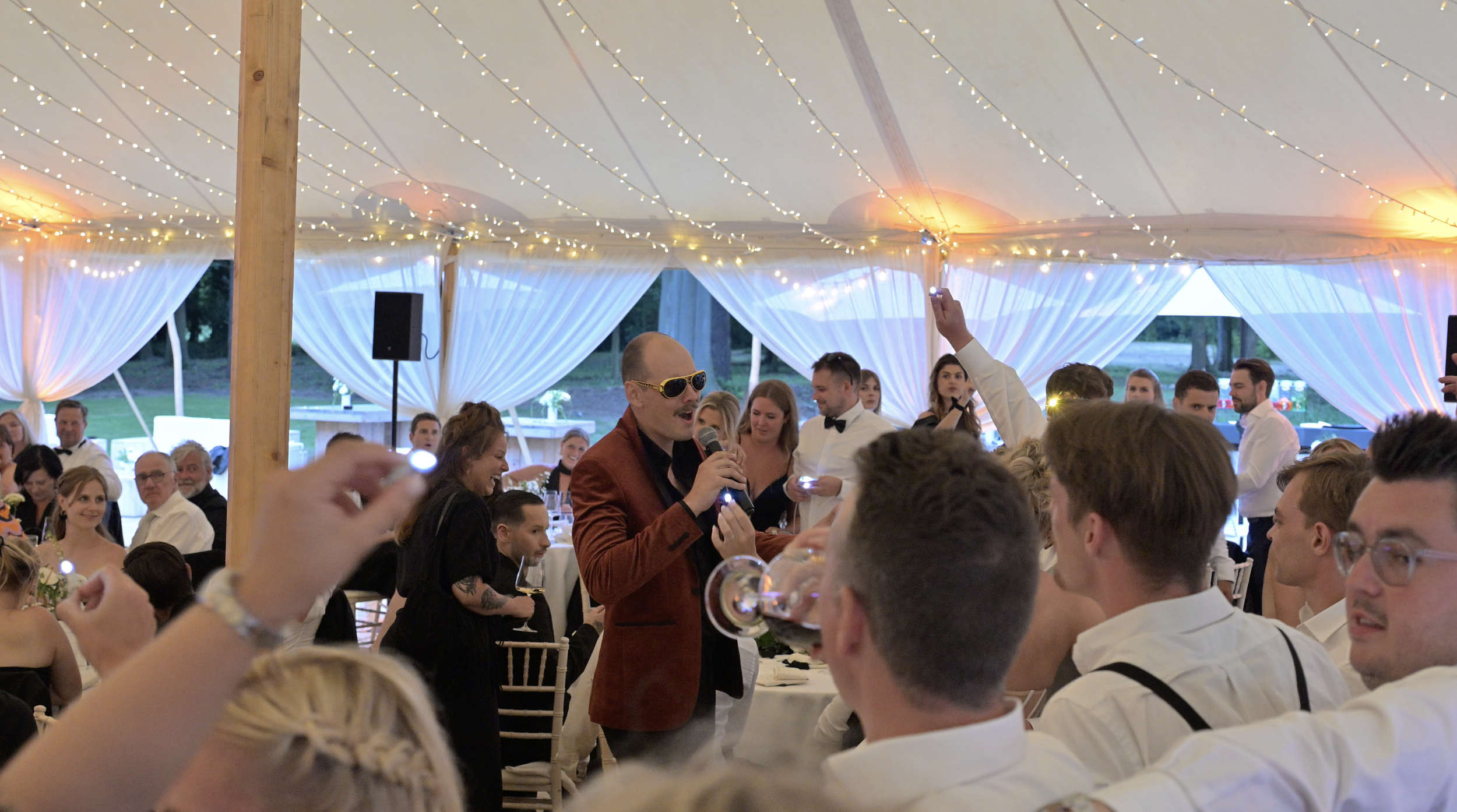 A man in a red suit jacket and sunglasses holding a microphone, speaking to a crowd at a wedding reception. The tent is decorated with string lights and white drapery.