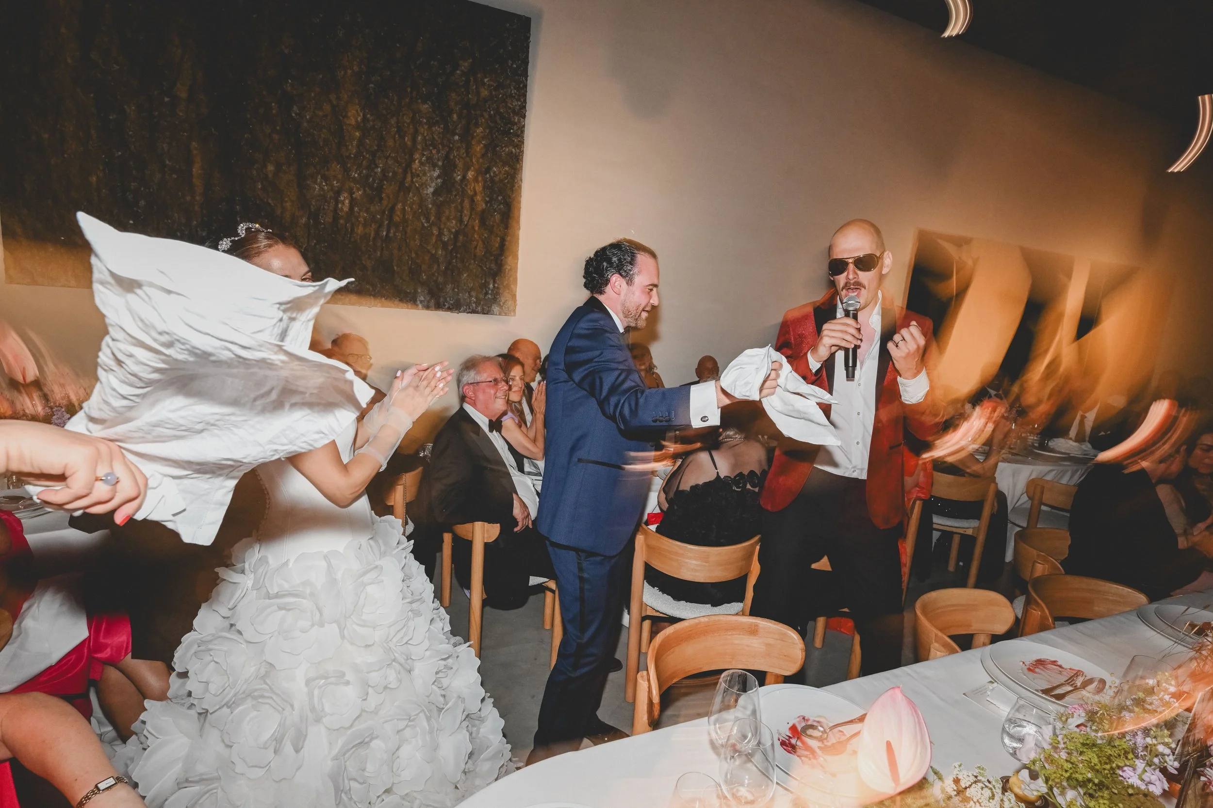 At a wedding reception, a woman in a wedding gown is clapping, and a man in a blue suit is smiling while holding a napkin. A man in a red jacket with sunglasses is speaking into a microphone. Other guests are seated at tables decorated with flowers and plates.