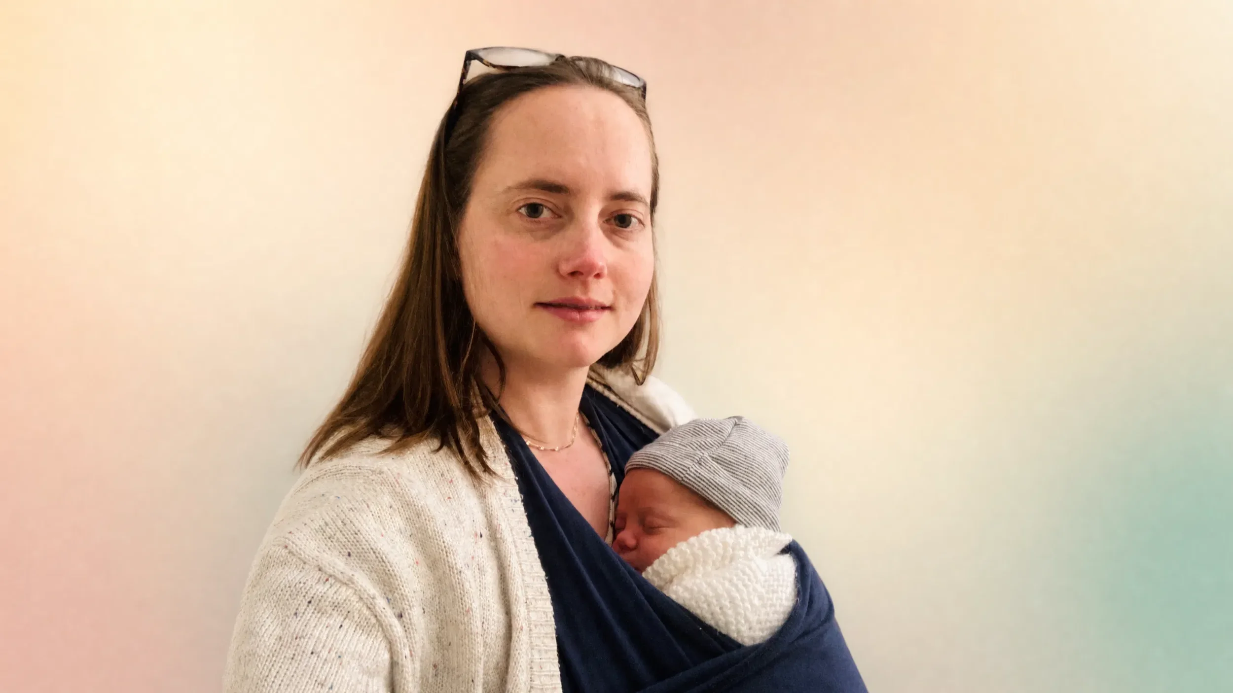 A woman with light skin and brown hair holding a sleeping baby wrapped in a blanket, standing against a plain light-colored wall. Signs of postpartum infection