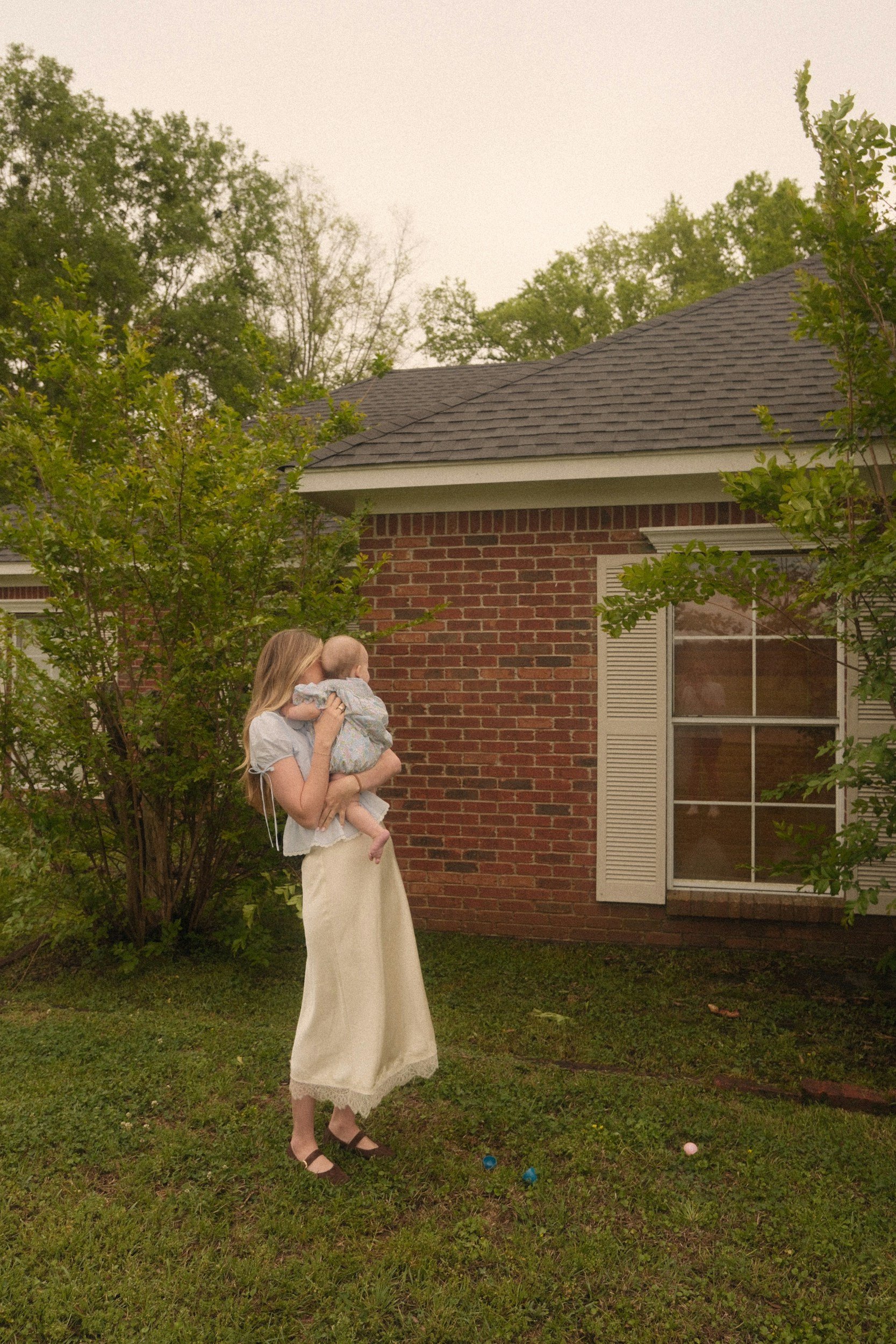 Woman in cream skirt and brown sandals holding a baby, standing on grass in front of a brick house with white window shutters, trees, and a cloudy sky.