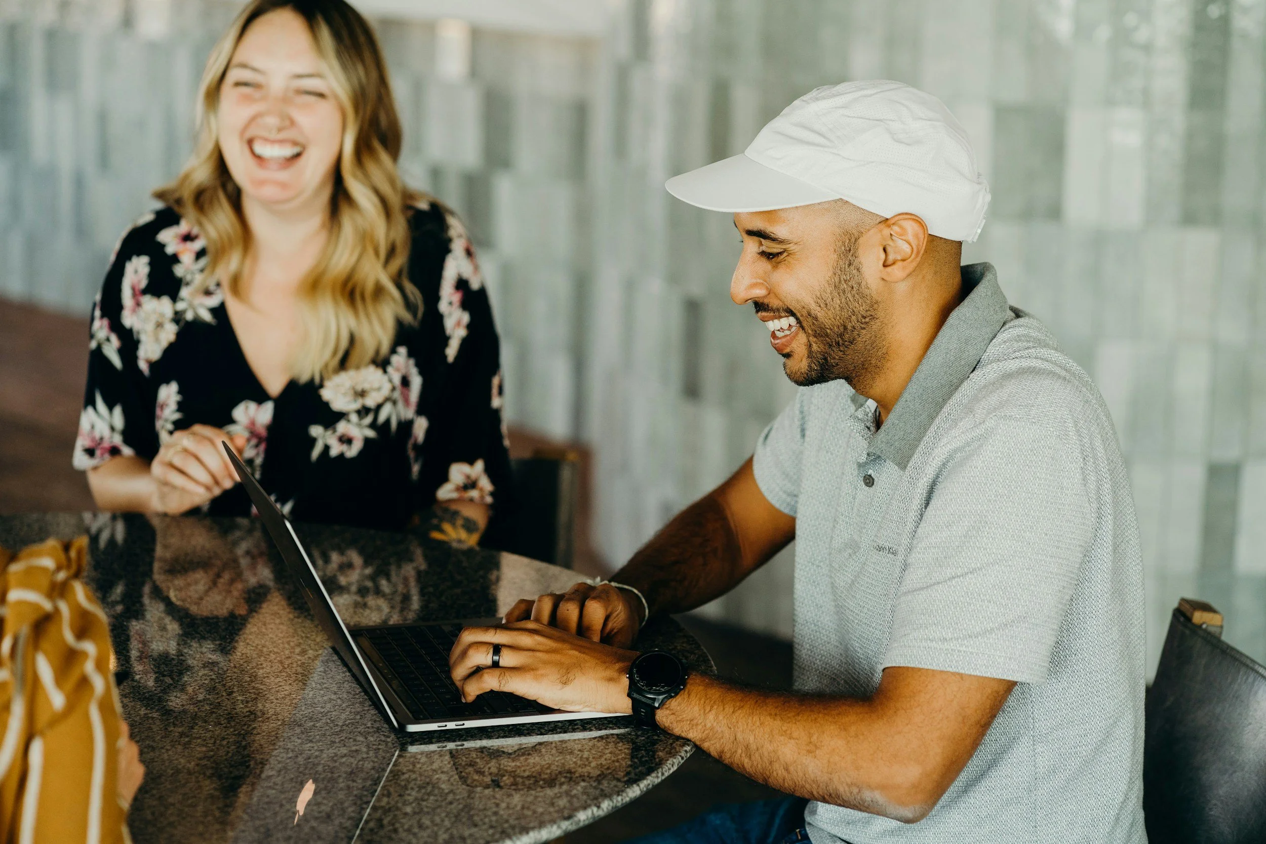 A man with a beard and a white cap laughing while working on a laptop at a kitchen counter with a woman in a floral dress standing next to him, also laughing.