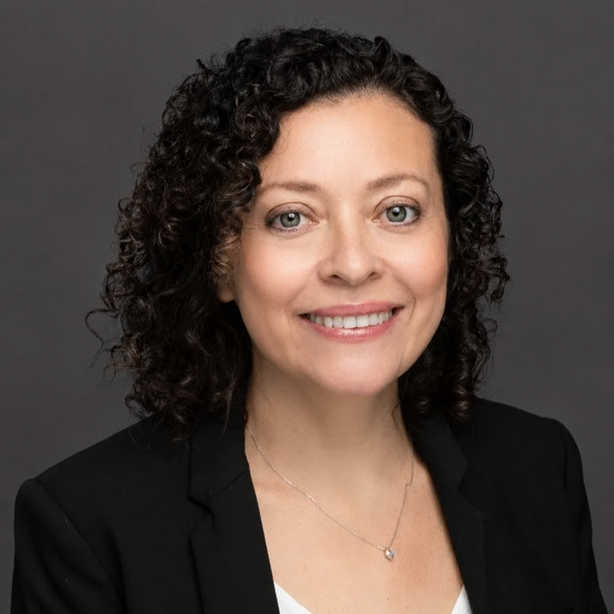 A portrait of a woman with curly dark hair smiling, wearing a black blazer and a necklace with a small pendant, against a gray background.