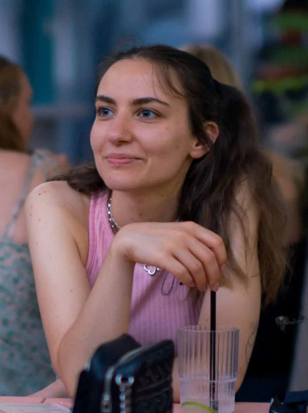 Junge Frau mit braunen, welligen Haaren und blauen Augen, die entspannt und glücklich in einem Café sitzt, trinkt ein Getränk mit Strohhalm, trägt eine rosa Top und eine Halskette.