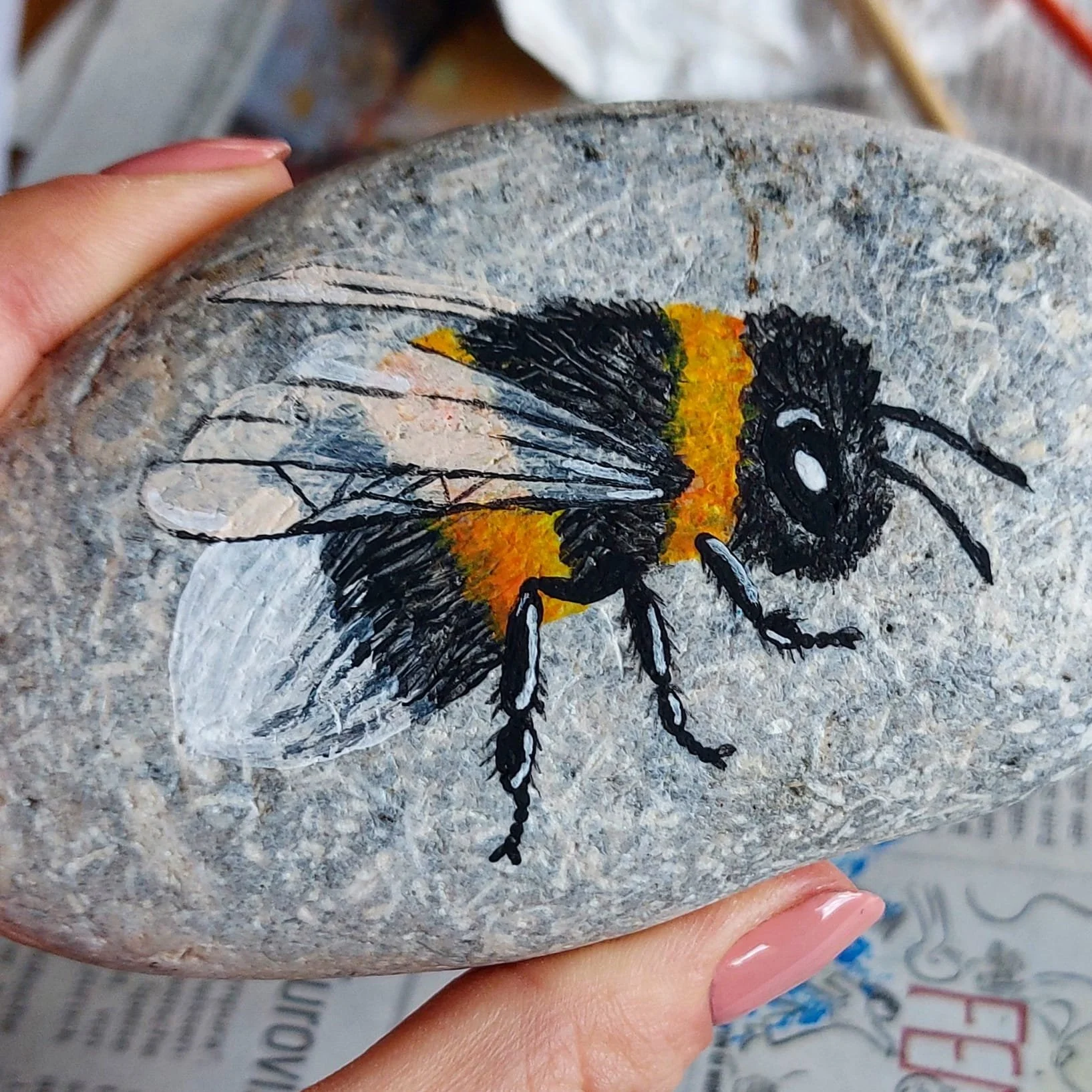 Eine bemalter Stein mit einem detailreichen Bienenbild, das eine Biene mit schwarzen und orangenen Farben zeigt, gehalten von einer Hand mit lackierten Nägeln.