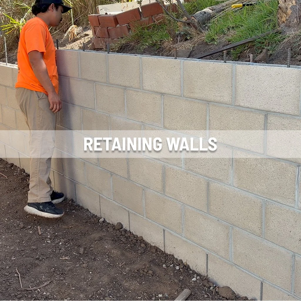 A man wearing an orange shirt and khaki pants standing next to a newly built concrete block retaining wall on a dirt ground. The wall has a steel reinforcement wire on top. Behind the wall, there are construction materials including bricks and soil.