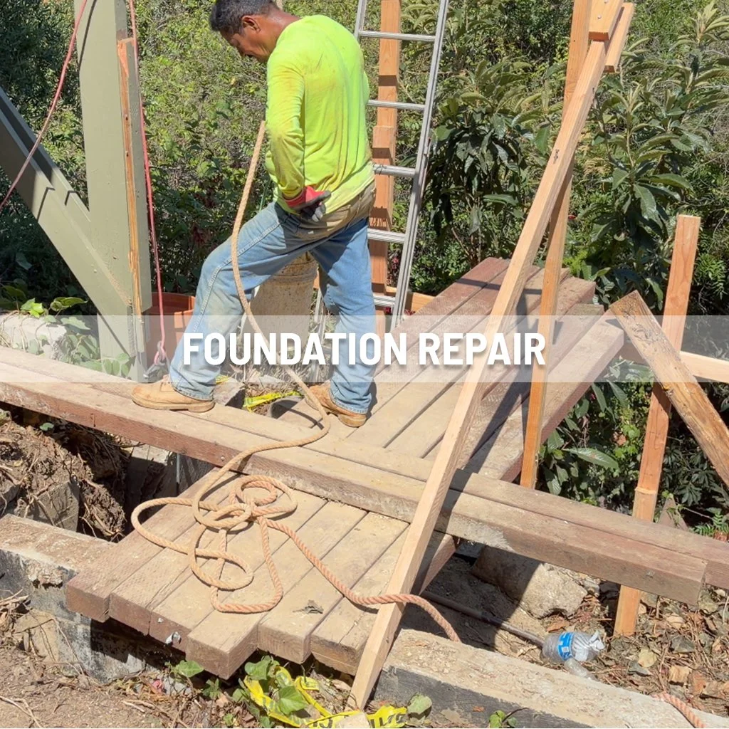 A worker in a neon yellow shirt and jeans repairs a building's foundation outdoors. He is standing on a wooden platform, surrounded by construction tools and materials, with trees in the background.