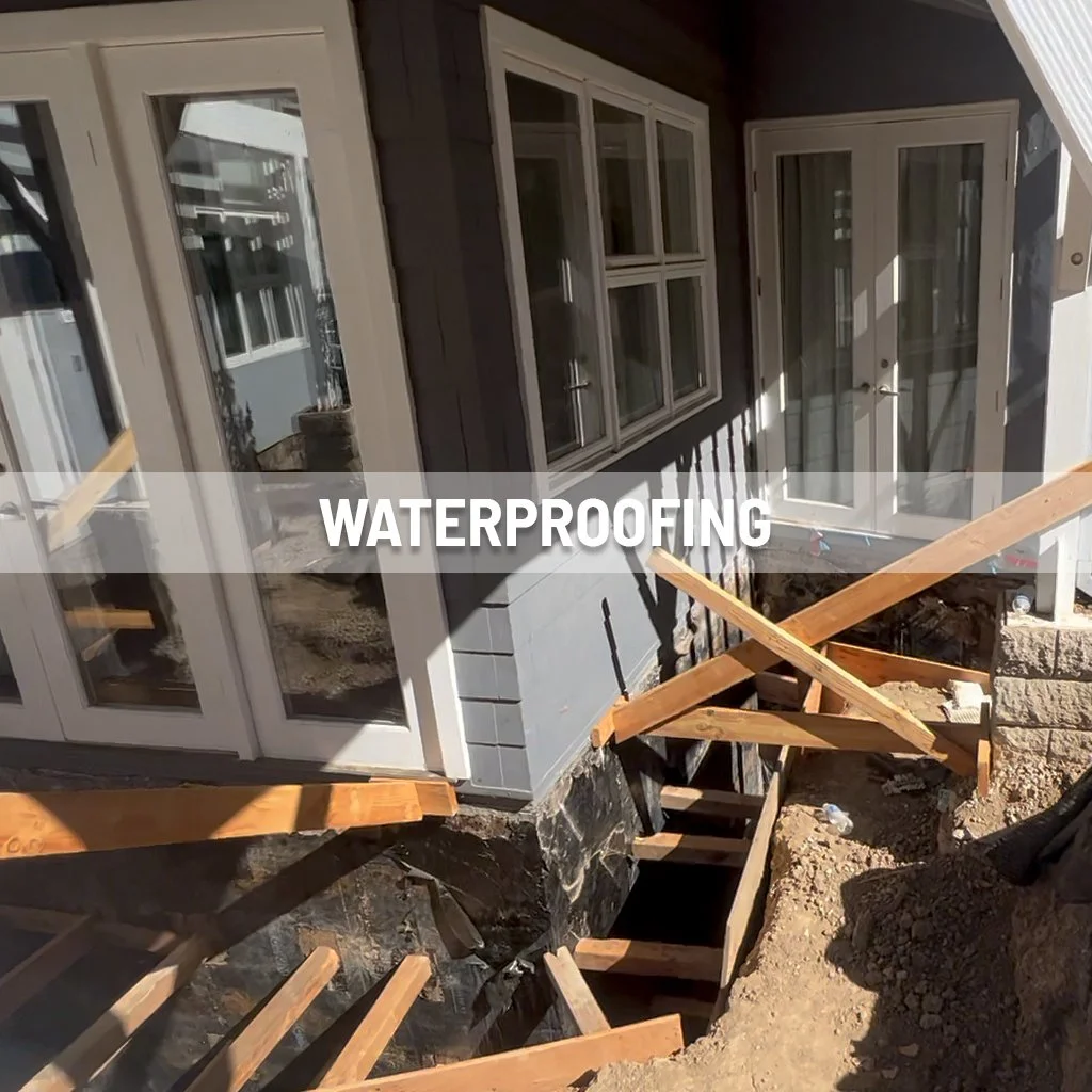 Construction site showing waterproofing work on the foundation of a house with wooden framing and a deck, with a door and windows installed.